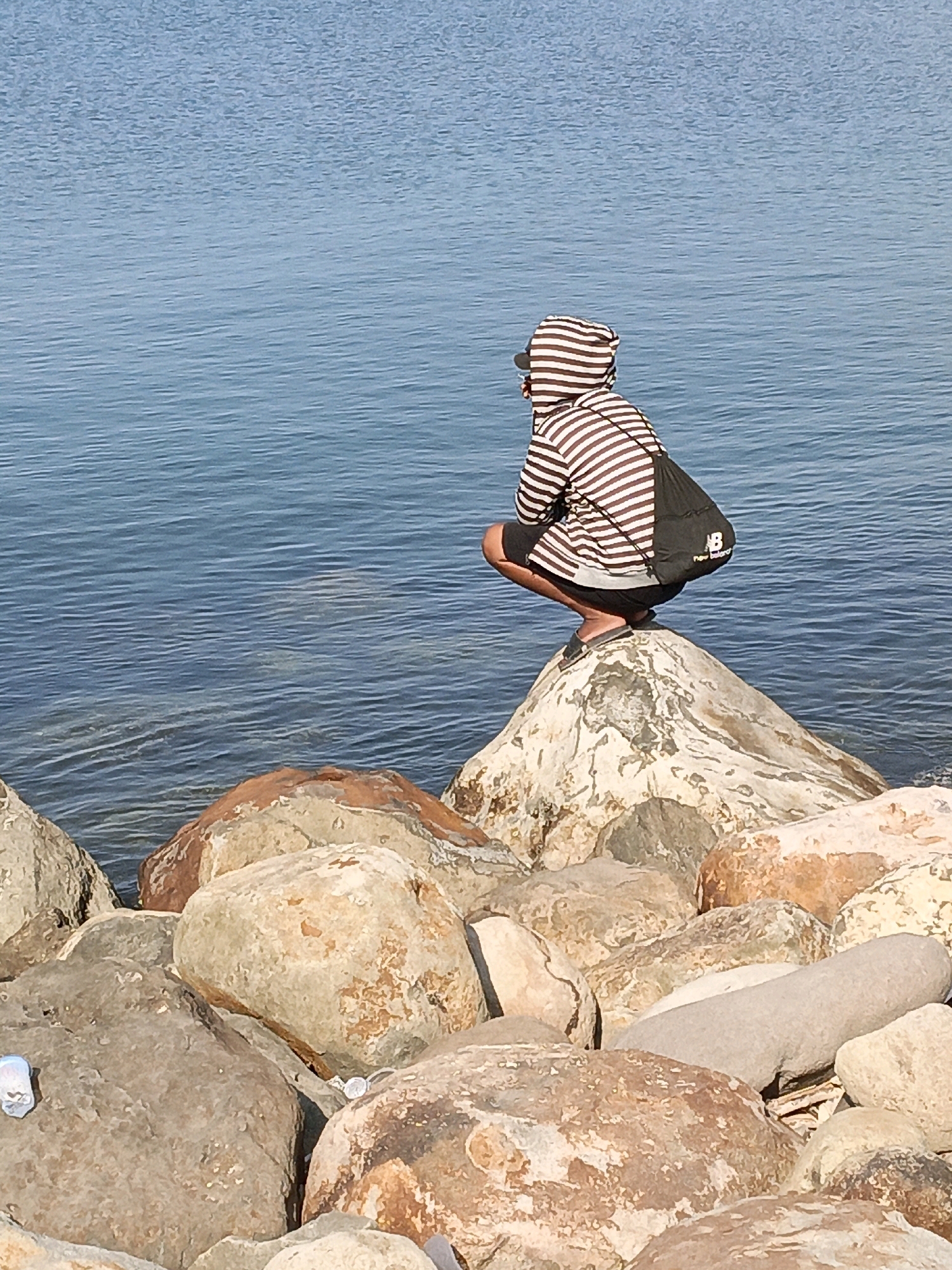 A person wearing a striped hoodie and black shorts crouches on a large, white rock. The person is looking out at the water. The water is blue and calm, with small ripples. The rocks in the foreground are various shades of brown and gray. A black drawstring bag with the New Balance logo is visible on the person's back. The image appears to be taken at a rocky shoreline. It is not possible to definitively identify the location as Bulukumba, Indonesia, based on the visual information provided in the image.