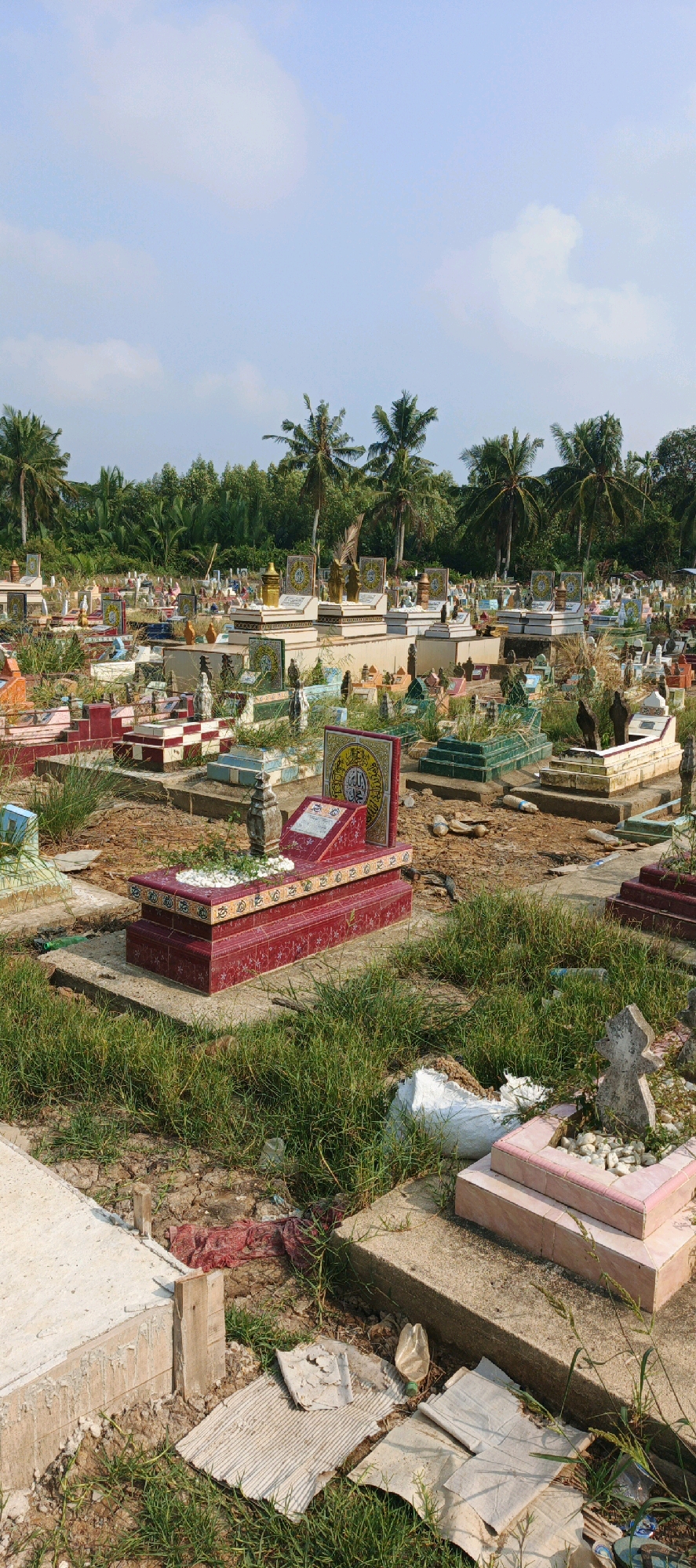 The image depicts a graveyard in Simbur Naik, Indonesia, under a clear sky with scattered clouds. The graveyard is densely populated with numerous tombs of varying sizes, shapes, and colors, including red, green, and white structures. Many of the tombs feature decorative tiles, some with Arabic inscriptions. Palm trees and other lush green vegetation surround the graveyard, suggesting a tropical or subtropical climate. The ground is covered with grass and dry earth, with some areas appearing unkempt. Several plastic bottles and pieces of debris are scattered amongst the tombs, indicating the presence of human activity and potentially a lack of regular maintenance. There are no people visible in the image. The scene appears to be an outdoor, established burial ground.