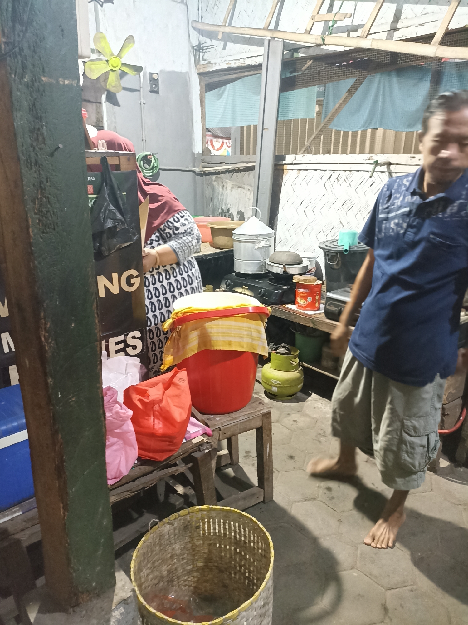 The image depicts an outdoor food stall or small eatery, likely in a residential or market area of Lumajang, Indonesia. A man in a blue polo shirt and light-colored shorts, barefoot, is walking away from the camera, his body blurred with motion. He appears to be a vendor or customer. Behind him, a woman wearing a headscarf and a patterned tunic is partially visible, interacting with the stall's setup.
The stall itself is a simple setup with various items for sale or preparation. There's a red plastic bucket with a yellow and white cloth cover, a green gas cylinder, and several cooking utensils on a stove. Bags, including a black plastic bag and a bright orange bag, are present, suggesting merchandise or ingredients. A yellow fan is mounted on a wall in the background, and the stall has a rustic roof structure made of bamboo. The ground is paved with hexagonal tiles. The lighting suggests it is either evening or a dimly lit indoor area. The overall impression is that of a casual, functional food service operation.