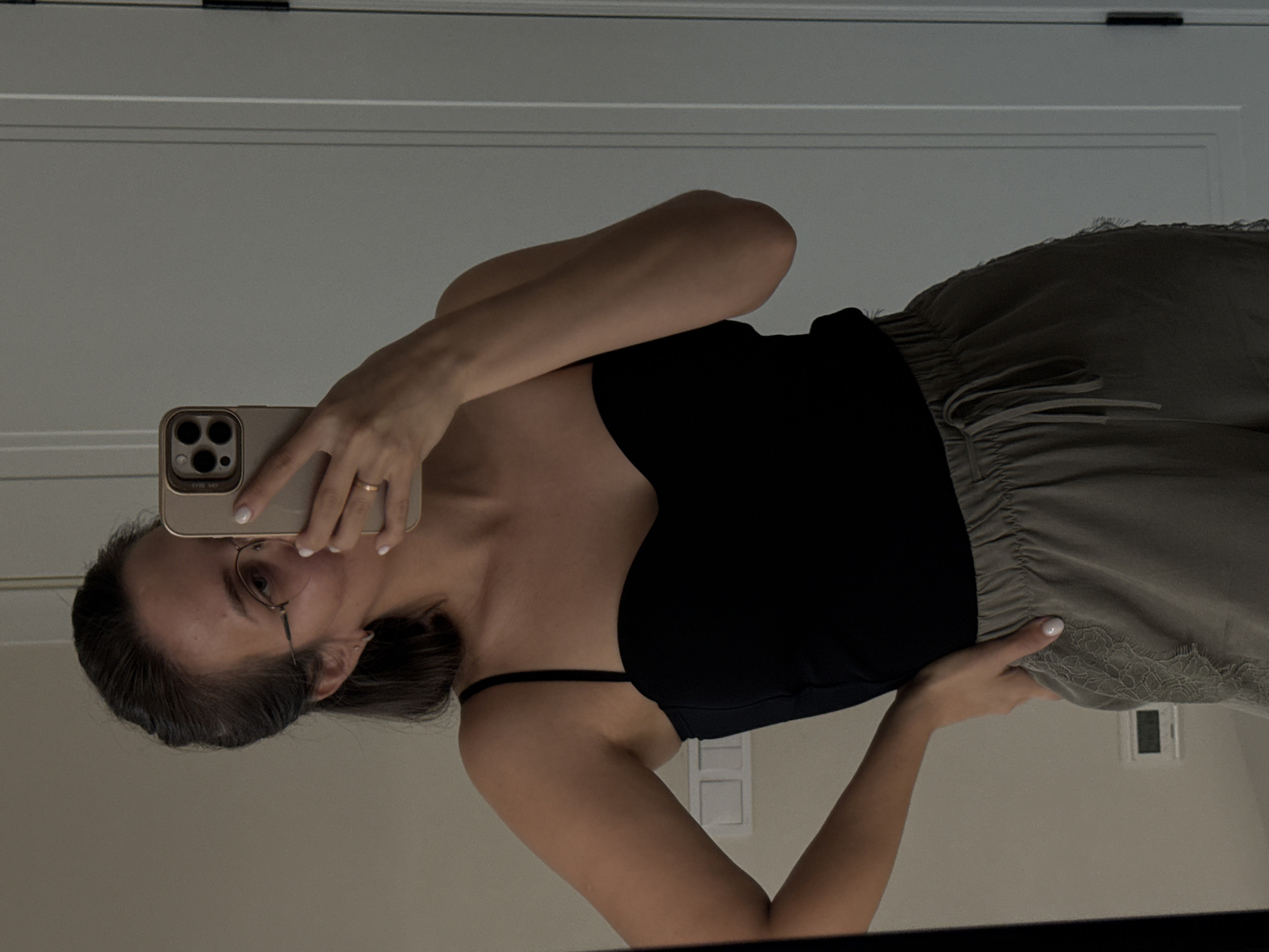 A woman is taking a mirror selfie in a well-lit room, possibly a bedroom or dressing area. She is wearing a black, strapless top with thin spaghetti straps and light-colored, drawstring pants that appear to have lace detailing along the side. She has her hair pulled back and is wearing glasses. Her right hand is on her hip, while her left hand holds a gold-colored smartphone, obscuring part of her face as she takes the photo. The background shows a light-colored wall with electrical outlets and what appears to be a white door on the right side. There is no indication of the city of Pervomaysky, Russia, in the image.