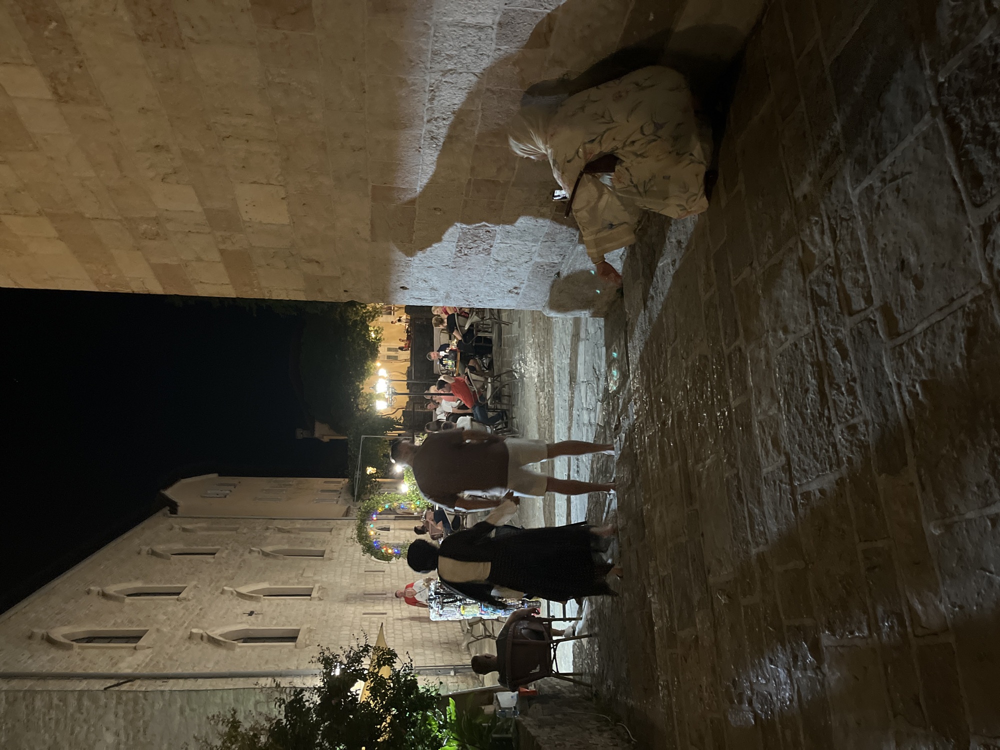 The image depicts a nighttime scene in an outdoor seating area of a restaurant or cafe, likely in the old town of Budva, Montenegro. The ground is paved with cobblestones that appear wet and reflective, suggesting recent rain or cleaning.
In the foreground, a woman wearing a white and floral patterned dress and a headscarf is sitting on a stone ledge against a textured stone wall. She is holding a mobile phone in her right hand, possibly taking a photo or recording. A dark bag is placed beside her.
Further back, a couple is walking away from the camera on the cobblestone path. The man is wearing a dark t-shirt and white shorts, and the woman is in a long black dress over a white garment.
To the left, a man is seated in a chair, facing away from the camera.
In the background, a busy outdoor dining area is visible. Several people are seated at tables, some of whom appear to be diners. String lights are hung above the seating area, providing illumination along with street lamps on poles. Buildings with arched windows line the street. The overall atmosphere is lively and communal.