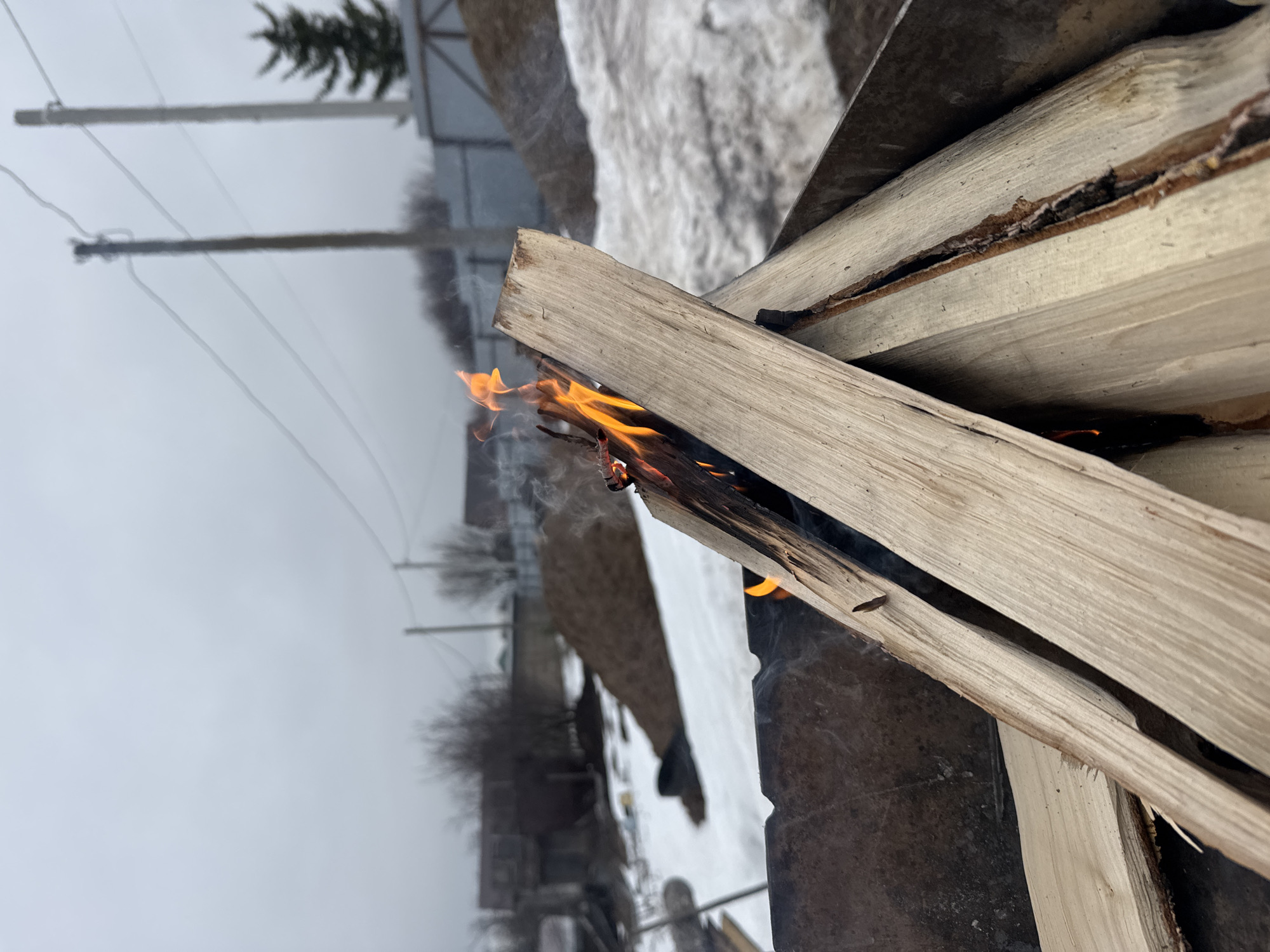 The image shows a small fire burning between stacked pieces of light-colored wood, likely firewood. The fire emits smoke. The wood is resting on a dark, possibly metal, surface. In the background, the scene is obscured by a light fog or overcast sky, revealing a rural or suburban environment. Bare trees, utility poles with wires, and a snowy patch are visible in the distance. Based on the prompt, this scene is assumed to be in Chaikovsky, Russia.