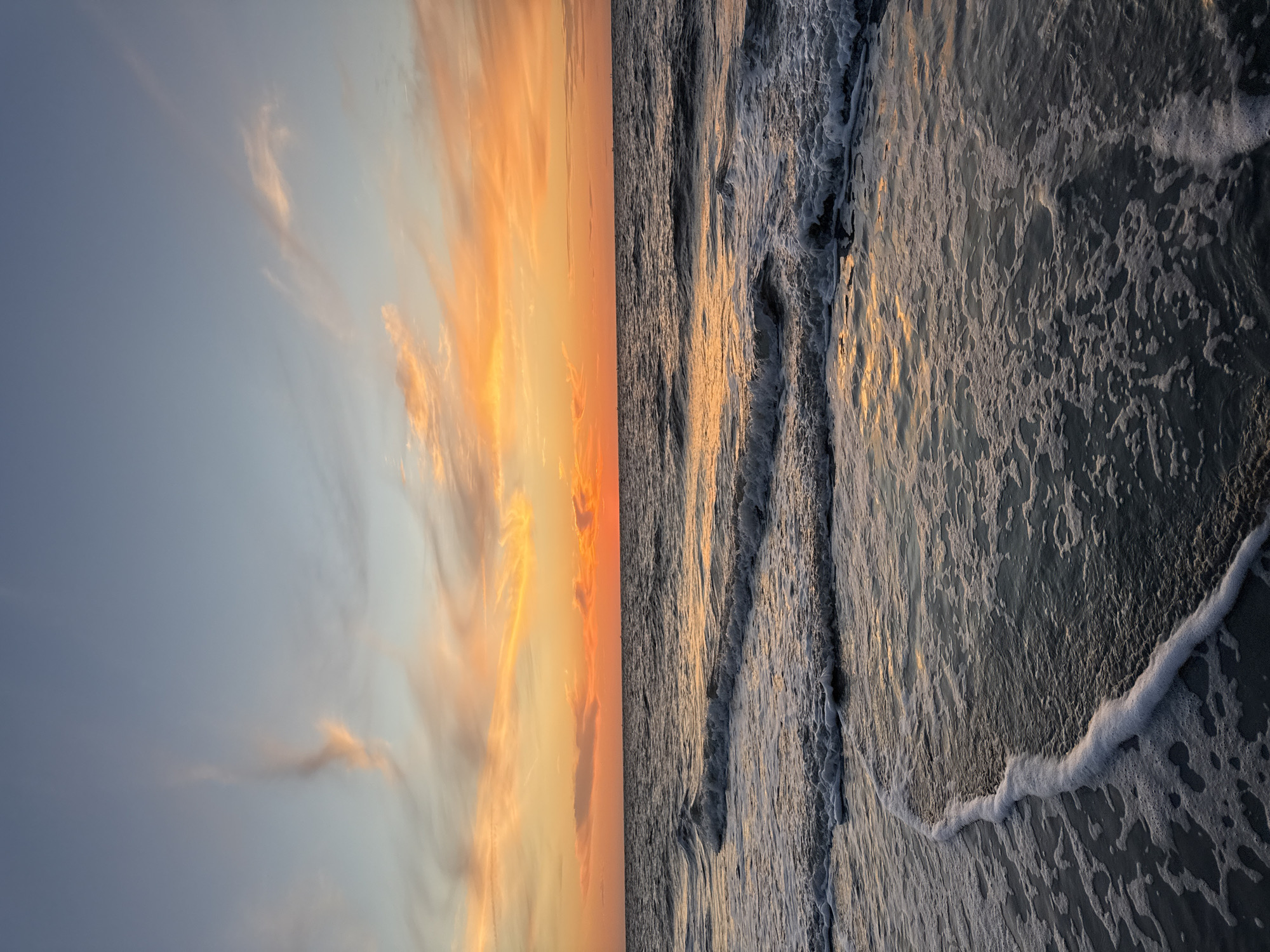 The image depicts a serene beach scene at sunset on the island of Langeoog, Germany. The sky is a gradient of soft blues and vibrant oranges, with wispy clouds catching the last rays of the sun. The horizon is marked by the calm sea, its surface reflecting the warm colors of the sky. Gentle waves are rolling onto the shore, creating a frothy white foam that trails along the wet sand. The overall impression is one of peacefulness and natural beauty, characteristic of a coastal sunset.