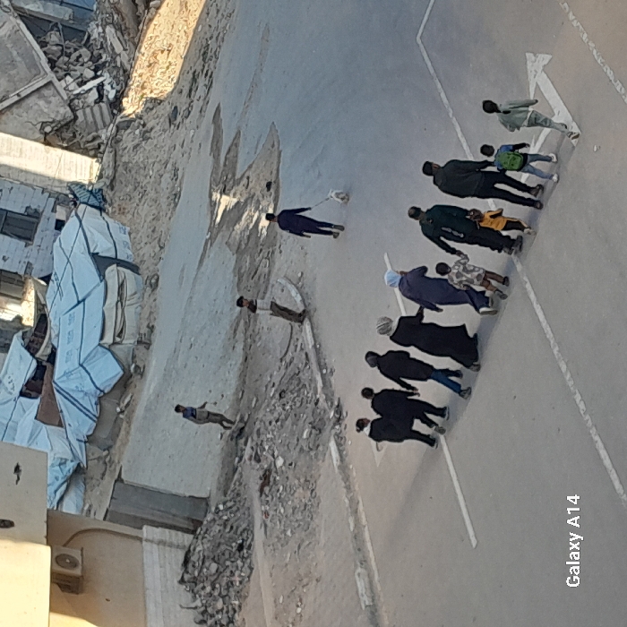 A group of adults and children are walking on a road next to a destroyed building and piles of relief supplies. The road has painted white lines dividing it. The people are mostly wearing dark-colored clothing, but some are wearing lighter shades. One person is walking a dog. The background shows signs of rubble and collapsed buildings. The image appears to be taken from an elevated position.