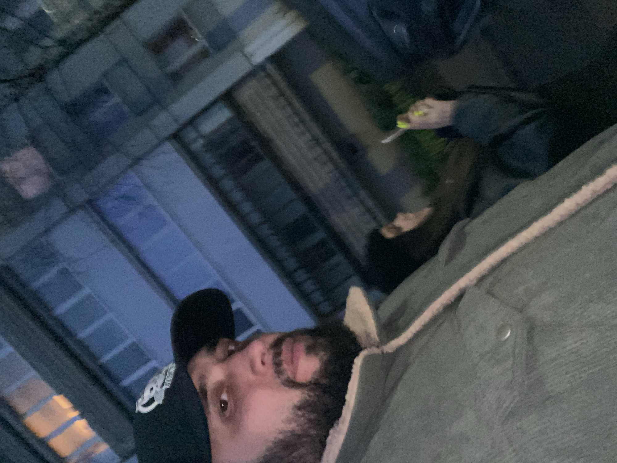 This media file depicts a casual, low-light scene captured at night or late dusk, likely in an urban area of Chaikovsky, Russia. The image is oriented sideways, with the main subject's head at the bottom.
In the foreground, a man with a beard and mustache is prominently featured, looking directly towards the camera with a neutral expression. He is wearing a dark baseball cap, which has an unreadable white logo or text on its front panel. He is also dressed in a dark grey or khaki jacket with a distinct light-colored, possibly fleecy, trim.
Further back in the mid-ground, on the upper right side of the frame (when correctly oriented), another individual is partially visible. Their hand and forearm are seen holding a slender white cylindrical object, possibly a cigarette or vape pen. A notable detail on this person is their bright, neon yellow or lime green painted fingernails.
The background consists of blurred, dark multi-story buildings with various windows, some reflecting ambient or artificial light. The overall lighting is dim, contributing to a blueish tint and a somewhat grainy quality throughout the image. The composition suggests a close-up, possibly selfie-style, photograph in an outdoor setting. No specific text beyond the unreadable logo on the cap is visible.