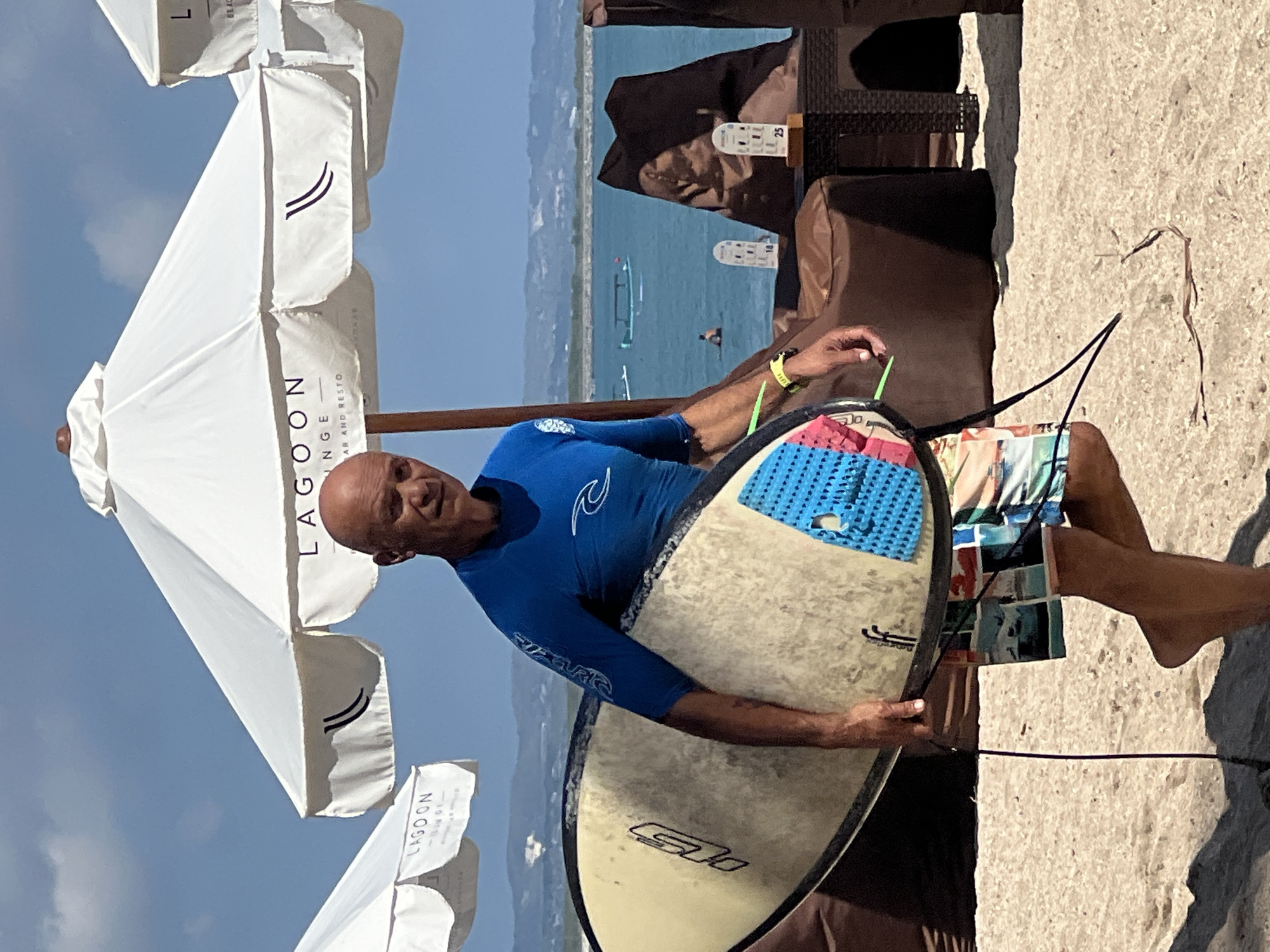 A bald, middle-aged Caucasian male wearing a blue rashguard and patterned swim trunks walks on a sandy beach carrying a surfboard. He is walking from left to right, facing the camera. The surfboard is off-white with black and blue accents. In the background, white beach umbrellas with the text "LAGOO N LOUNGE" are visible. The ocean is in the distance with a few boats and people swimming. The setting appears to be a beach resort in Kuta, Indonesia, given the distinctive beach umbrellas and the presence of traditional Indonesian outrigger boats in the water. The day is sunny with a clear blue sky.
