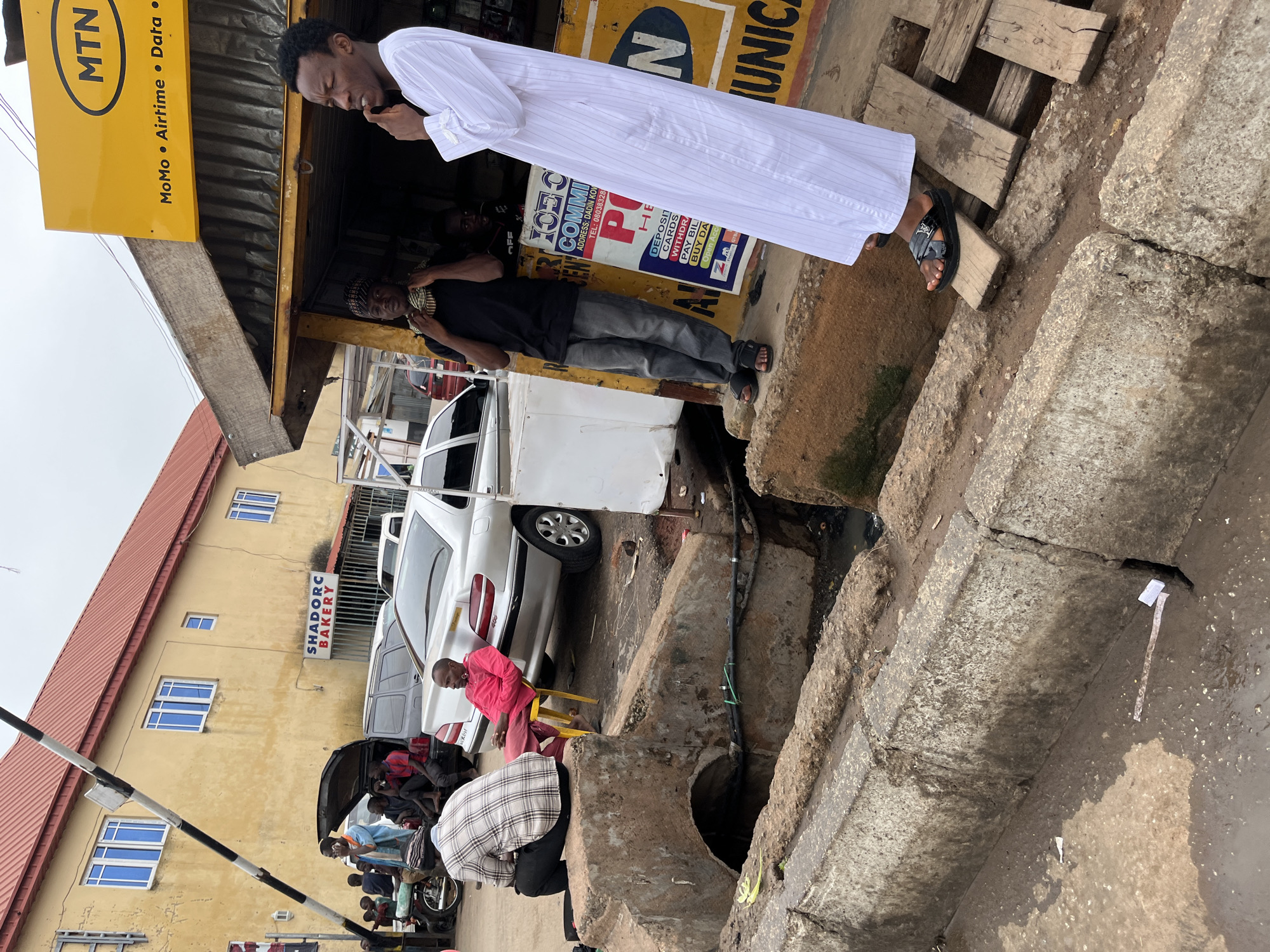This image captures a bustling street scene in Gura Top, Nigeria, featuring multiple people, commercial establishments, and everyday urban elements. The photograph is taken at an angle, giving a slightly tilted perspective of the street and buildings.
In the foreground, to the right, a man wearing a long white traditional garment (possibly an agbada) and sandals stands near a storefront, looking downwards or to the side. Behind him, closer to the shop entrance, another man in a black t-shirt and grey trousers appears to be adjusting something around his neck. Further back, inside the shop, other indistinct figures are visible. The ground directly in front of them features an open concrete drainage ditch, spanned by several wooden planks serving as a makeshift bridge. A black pipe runs inside the ditch.
To the left and further down the street, a multi-story building with a red roof and yellow walls, featuring blue-framed windows, is visible. A sign on this building reads "SHADORC BAKERY." In the mid-ground, a white car, resembling a Peugeot 406, is parked. Nearby, a man in a red shirt and light trousers sits on a yellow stool, while another individual in a plaid shirt bends over near the ditch. Towards the bottom left, a black and white striped barrier pole is present, and several people are gathered around what appears to be a tricycle (Keke Napep).
Visible text on the prominent yellow sign identifies it as an "MTN" service point, offering "MoMo • Airtime • Data •". Below this, another sign for a "COMMUNICATIONS" shop lists services like "DEPOSIT CARDS," "WITHDRAW," "PAY BILL," and "BUY DATA," along with an "ADDRESS: DADIN KOV" and a phone number "TEL: 08038328".
The scene suggests a typical daytime setting, with an overcast sky visible at the top, indicating a cloudy day. No strong emotions are apparent, as people seem engaged in routine activities, possibly transacting business at the shops or moving through the street. The visible infrastructure, including the open drainage and makeshift bridge, points to the local urban environment of Gura Top.