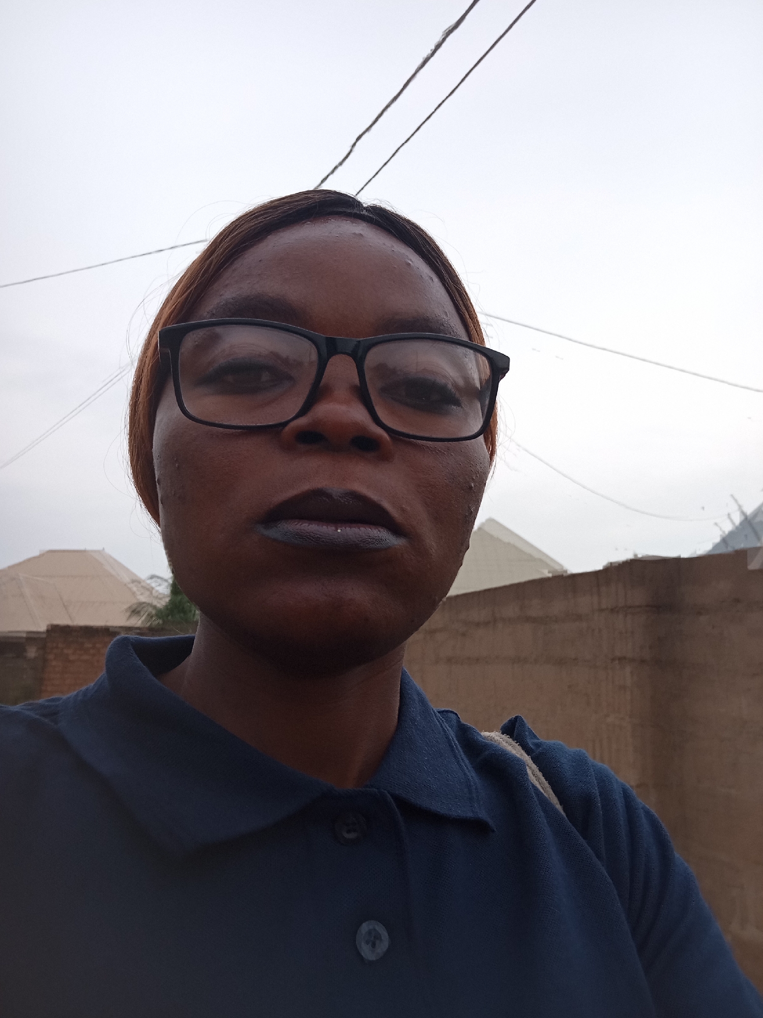 A close-up, selfie-style shot features a person with dark skin and brown hair, wearing black-rimmed glasses and a dark blue collared shirt. Their lips are painted with dark lipstick. The background appears to be outdoors, with a light-colored, overcast sky above. In the middle ground, there is a muted brown wall and what appear to be parts of buildings with peaked roofs. The overall impression is a casual, everyday moment captured in an urban or semi-urban setting. The image is likely taken in Kason, Nigeria, given the context provided.
