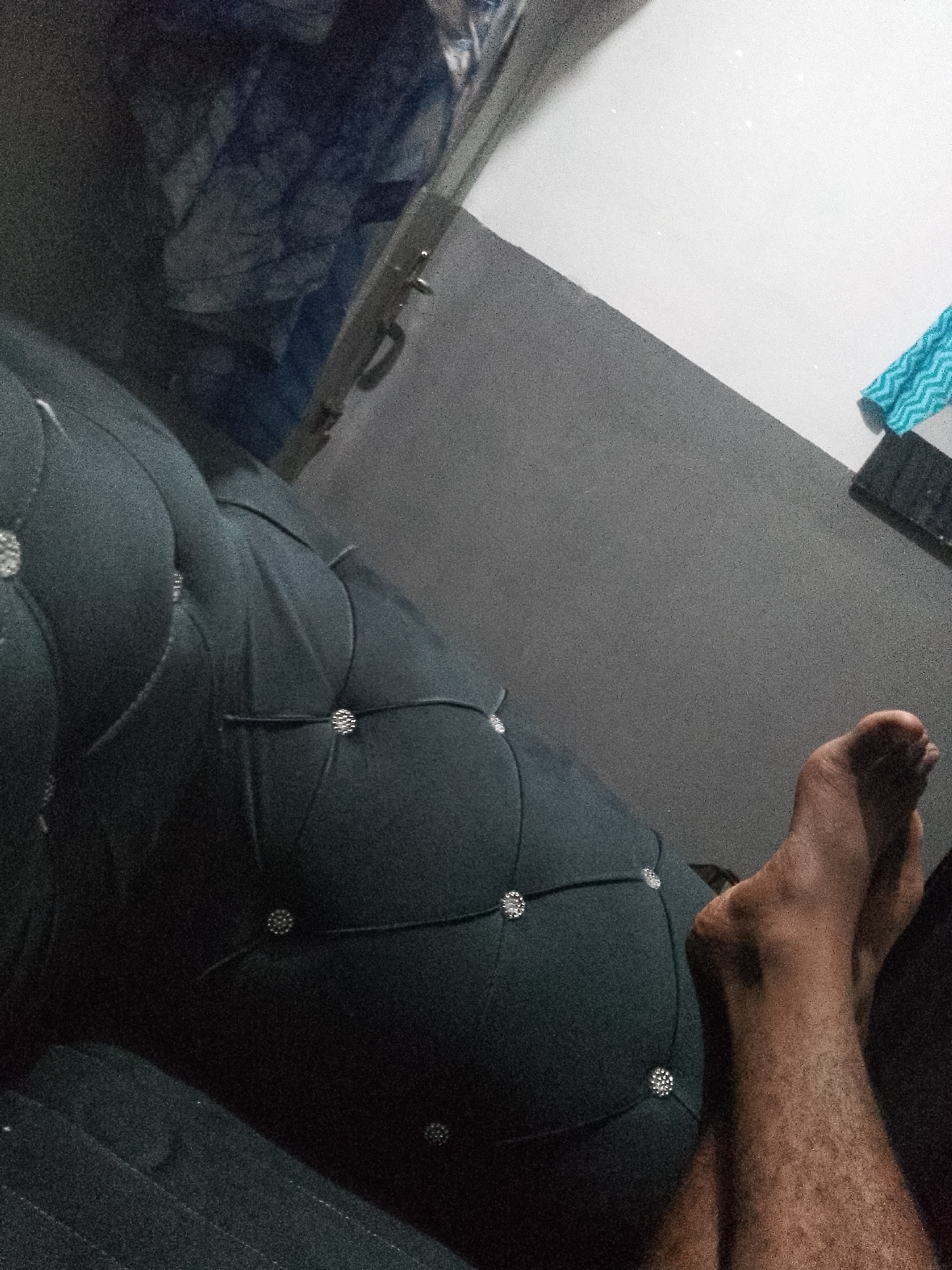 The image depicts a close-up view of a person's foot resting on a dark, tufted surface, possibly a piece of furniture. The foot is bare and appears to be brown in skin tone, with visible hair on the leg. The surface it rests on is dark and appears to have a diamond-patterned quilting with decorative studs. In the background, the environment is dimly lit and appears to be indoors, with a gray wall and a lighter colored surface that may be a table or counter. A blue and white patterned cloth is visible in the upper right corner. The overall scene is grainy and lacks clear detail of the surroundings. There is no information or visual cue in the image to suggest a location in Jauro Yinu, Nigeria.