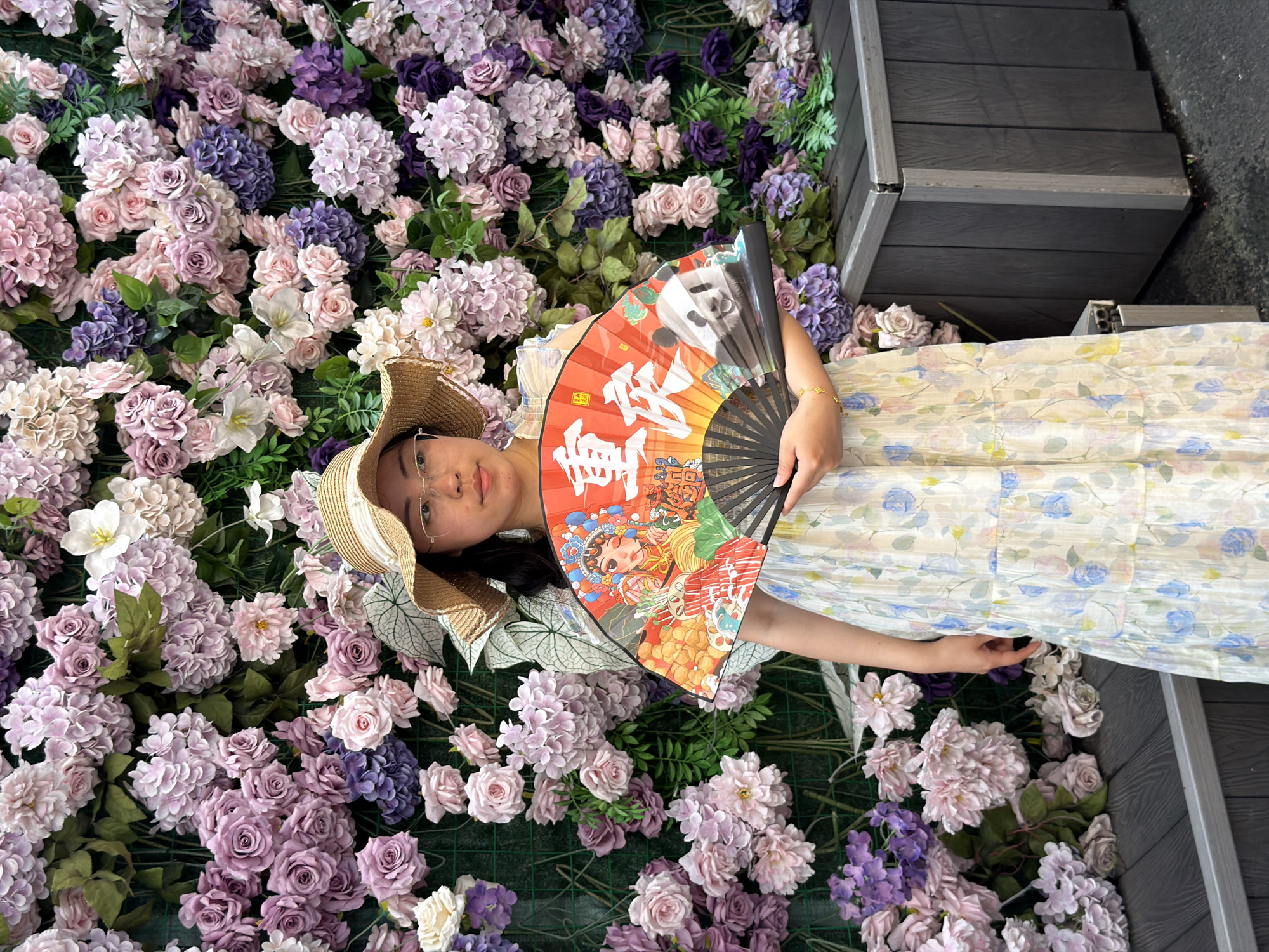 A young East Asian woman wearing glasses and a straw hat stands in front of a wall of artificial flowers in a scene located in the Yuzhong District of China. She is holding an open fan decorated with an image of Peking Opera performer and a panda. The woman is wearing a white, tiered dress with a floral pattern of blue and yellow. The flower wall behind her is a dense arrangement of various shades of purple, pink, and white flowers with green leaves. In the foreground, there is a dark gray wooden planter box and a wooden platform.