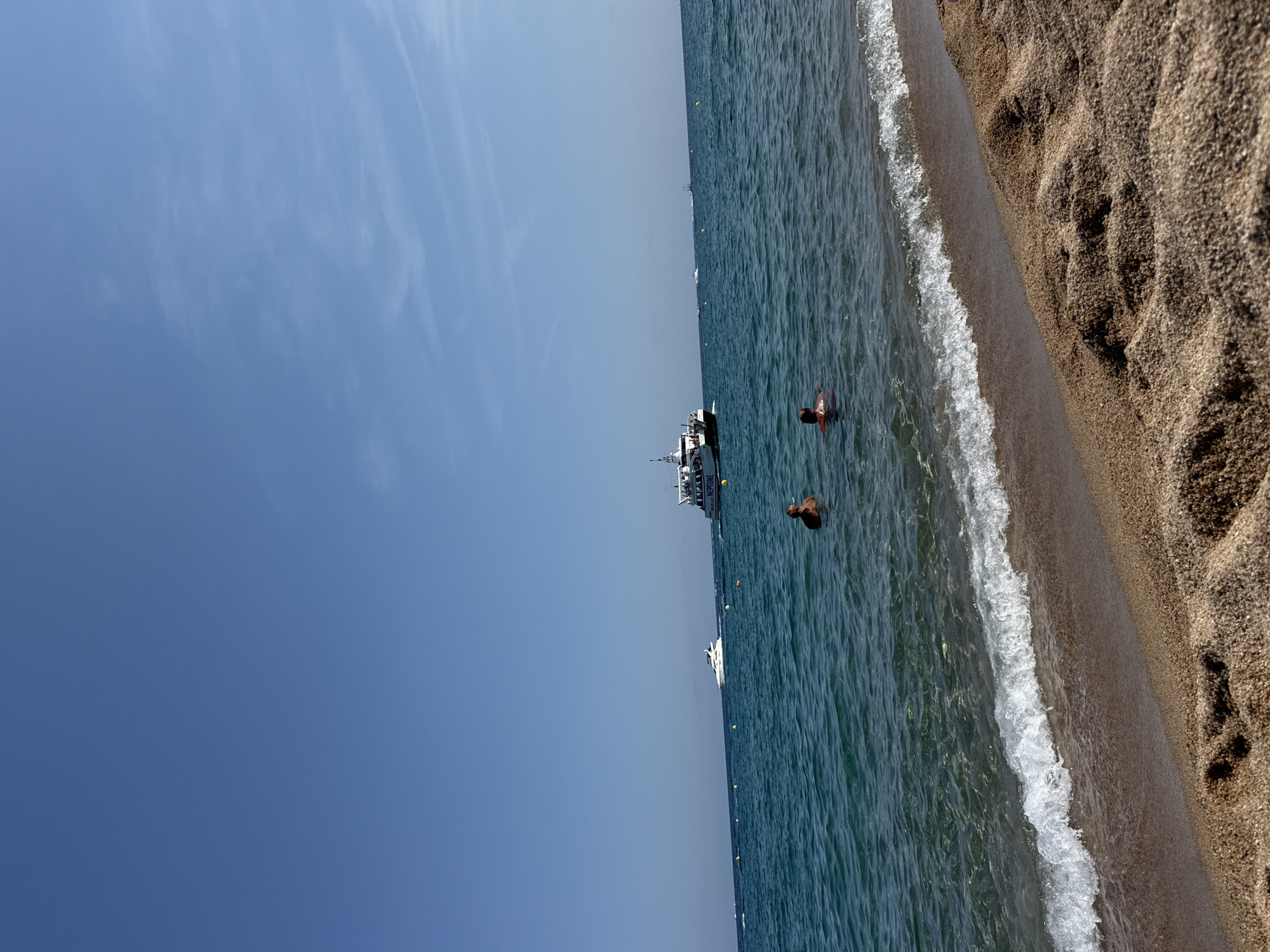 The image depicts a sunny day at a beach in Blanes, Spain. In the foreground, a sandy beach is visible with gentle waves lapping at the shore. Further out in the clear turquoise water, two people are swimming. One person is closer to the shore and appears to be a man, facing away from the camera. Another person is further out, also facing away. In the middle ground, several boats are present. One is a larger white ferry-like vessel with passengers visible on its deck, and the name "NEPTUNE" is visible on its side. Another, smaller white yacht is also visible in the distance. Yellow buoys mark an area in the water. The sky is a clear, bright blue with a few wispy clouds. The scene suggests a leisurely summer day at the coast.