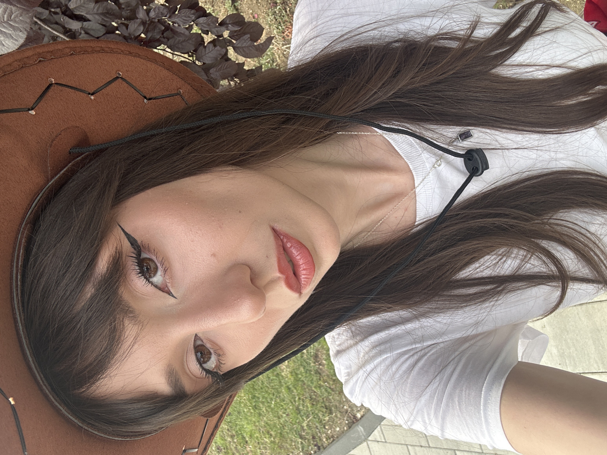 In a close-up, outdoor, portrait-style photograph, a young woman with long, dark brown hair and bangs is wearing a brown cowboy hat and a white t-shirt. She is positioned slightly off-center to the left, with her head tilted and looking directly at the camera. Her eyes are accentuated with black winged eyeliner and mascara. She is wearing lip gloss and has a faint blush on her cheeks. A thin silver chain necklace with a small pendant is visible around her neck, and she is also wearing a lanyard with a black adjuster and a small purple pendant. The background consists of blurred greenery and dark foliage, suggesting an outdoor setting, possibly a park or garden. There is no indication of the specific location within Grocka Urban Municipality, Serbia.
