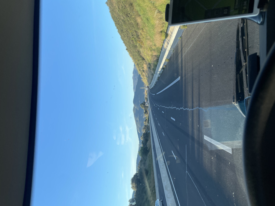 The image presents a first-person view from inside a vehicle, likely a truck, traveling on a multi-lane asphalt highway near Tresserre, France. The road ahead curves slightly to the left, marked with white dashed and solid lane lines, and shows a distinct crack running through the current lane's asphalt. To the left, a concrete barrier separates the highway from a parallel road where a light-colored sedan is visible, followed by a landscape of green vegetation, trees, and distant hills under a clear blue sky. To the right, a steep embankment covered in green and dry vegetation rises, bordered by a concrete safety barrier. Further down the highway, a white truck and other vehicles are discernable, along with a metallic power line pylon on the right side. The scene is illuminated by direct sunlight, casting long shadows across the roadway. In the immediate foreground, the top rim of a steering wheel is visible, and on the lower right, a dashboard-mounted navigation device displays a green map interface. The vehicle's windshield wipers are present at the bottom of the viewable area.