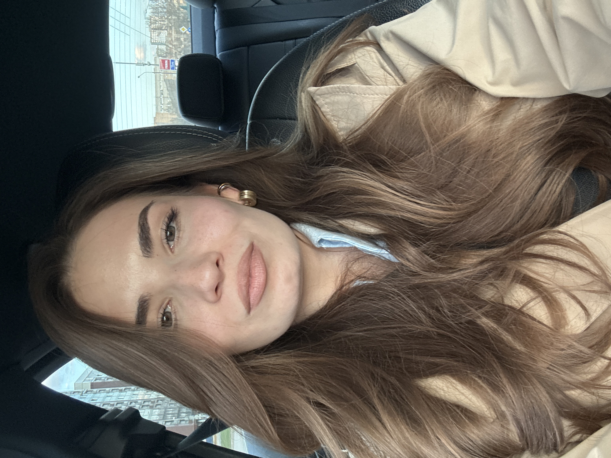 A woman with long brown hair sits in the driver's seat of a car. She is looking directly at the camera. She wears a light-colored coat. Outside the car window are buildings, indicating an urban environment that is Moscow, Russia. A gas station is visible in the background.