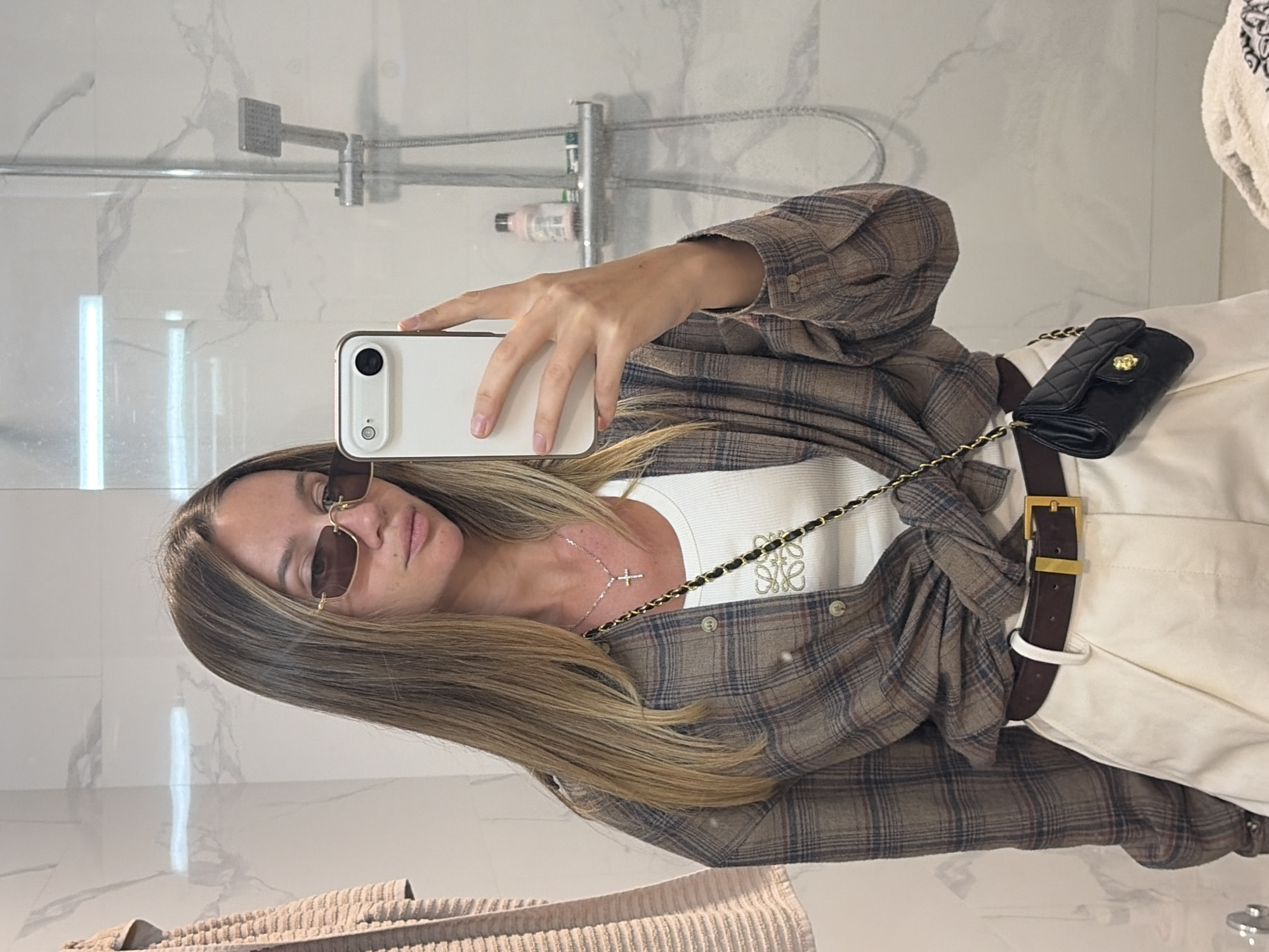 A person is taking a selfie in a bathroom setting. The individual is wearing sunglasses, a white tank top with a logo, a plaid shirt, white pants, and a brown belt. A black purse with a gold chain is visible. The bathroom features a showerhead, a white marble wall, and a beige towel.