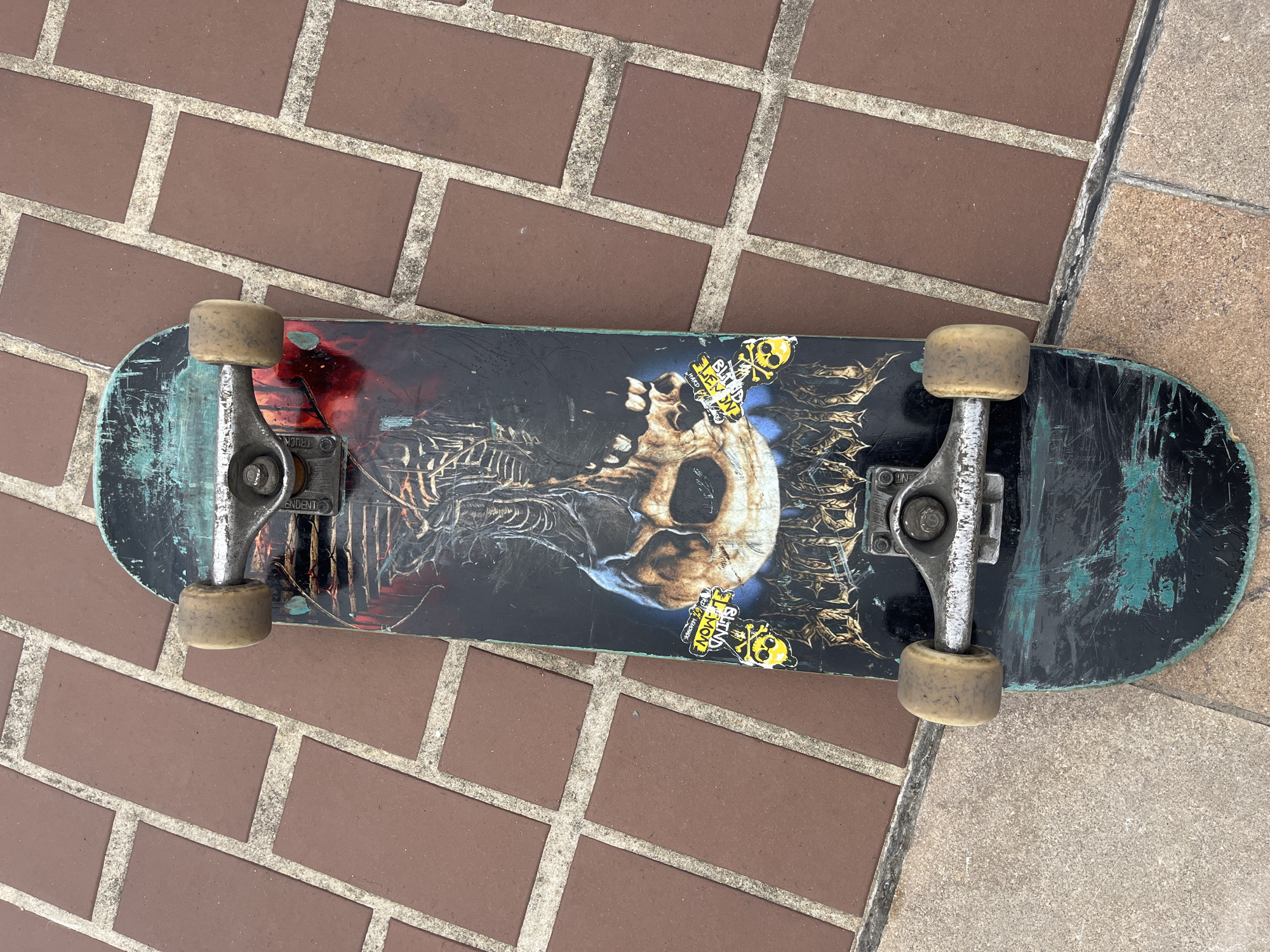 A black skateboard with a graphic of a skeleton and skull is lying on a brick-patterned sidewalk. The skateboard has two silver trucks and four yellowed wheels. The graphic on the skateboard features a skeletal ribcage with a skull at the center. There are also some stickers on the skateboard. The sidewalk is made of rectangular red bricks with white grout lines. The scene is outdoors, and the lighting suggests it is daytime. There are no people visible in the image. The skateboard appears worn, with scuff marks and faded paint, indicating it has been used. The overall environment suggests a casual urban setting. The location is the city of Créteil, France.
