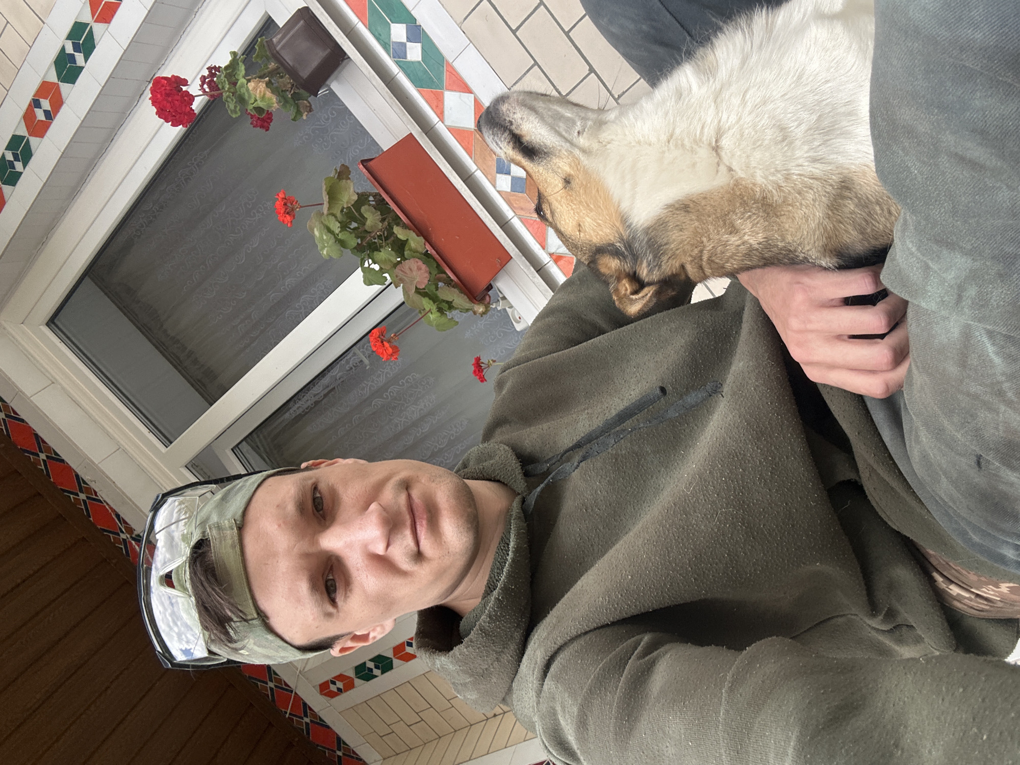 A man wearing a green camouflage baseball cap with clear safety glasses on top and a green hooded sweatshirt is sitting on an outdoor porch. He is looking towards the camera with a neutral expression. Next to him, a dog with tan and white fur is resting its head on his lap. The dog is looking upwards, away from the camera. Behind them, a window with white trim is visible, with red geraniums in brown planters on the windowsill. The porch has decorative tiles on the wall. The location is identified as Makiv, Ukraine.