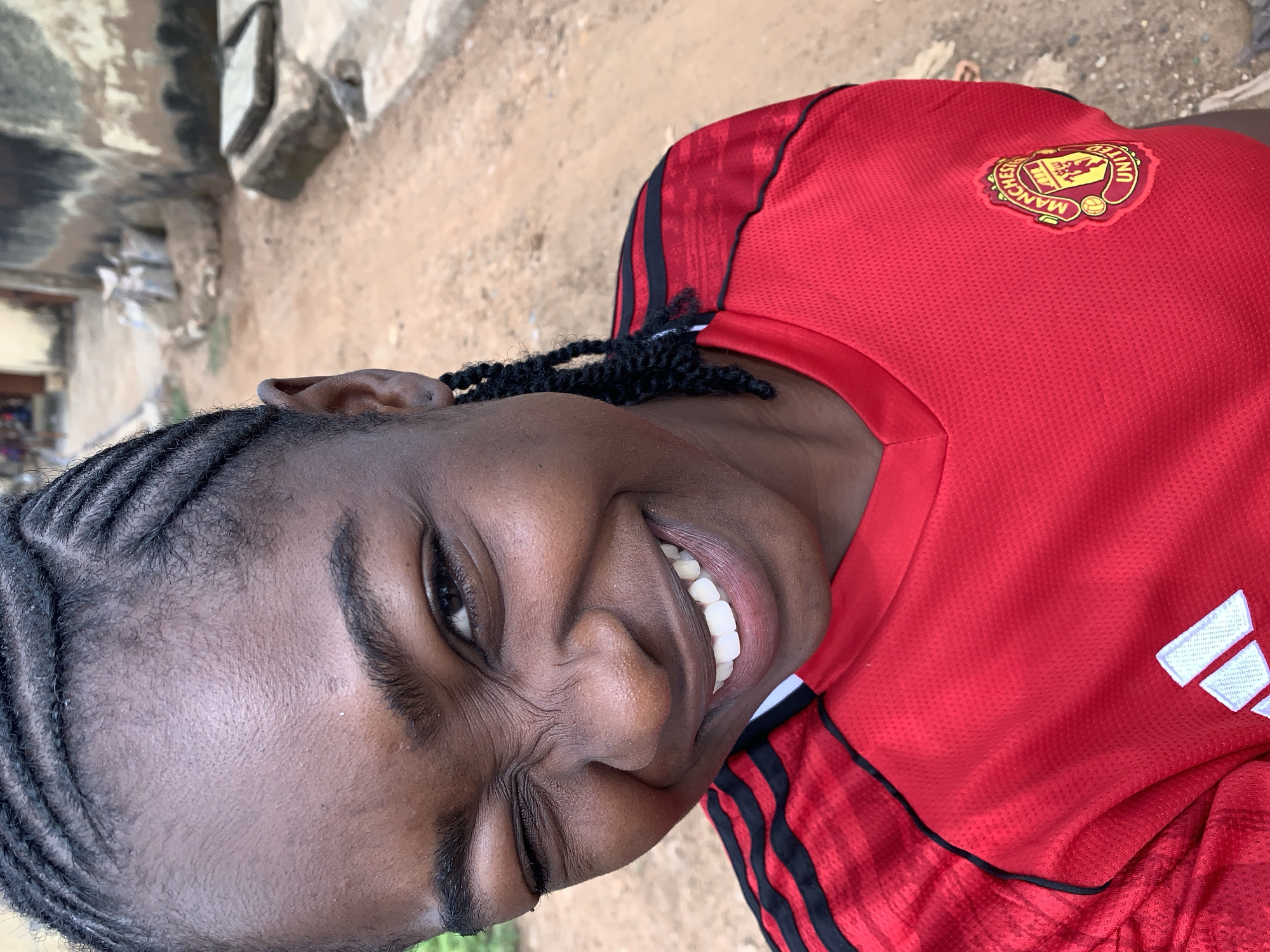 The image features a young person with dark skin, positioned in a half-profile and smiling broadly directly at the viewer. They have intricately braided dark hair, styled in cornrows across their scalp. The person is wearing a red short-sleeved sports jersey with black stripes on the shoulders and black trim.
On the left chest of the jersey, the Manchester United Football Club crest is prominently displayed. The crest is primarily red, black, and yellow, and features a devil mascot, a ship, and three roses, encircled by the text "MANCHESTER UNITED" and "FOOTBALL CLUB." On the opposite side of the chest, a white Adidas logo, characterized by three parallel bars, is visible.
The background is softly blurred, indicating an outdoor setting during the daytime. The ground visible behind the person appears to be unpaved, consisting of light brown earth or sand. Some indistinct structures or debris are faintly visible in the far background. The person's wide smile and crinkled eyes convey a strong sense of happiness and joy. This scene is set in Rafin Par, Nigeria.