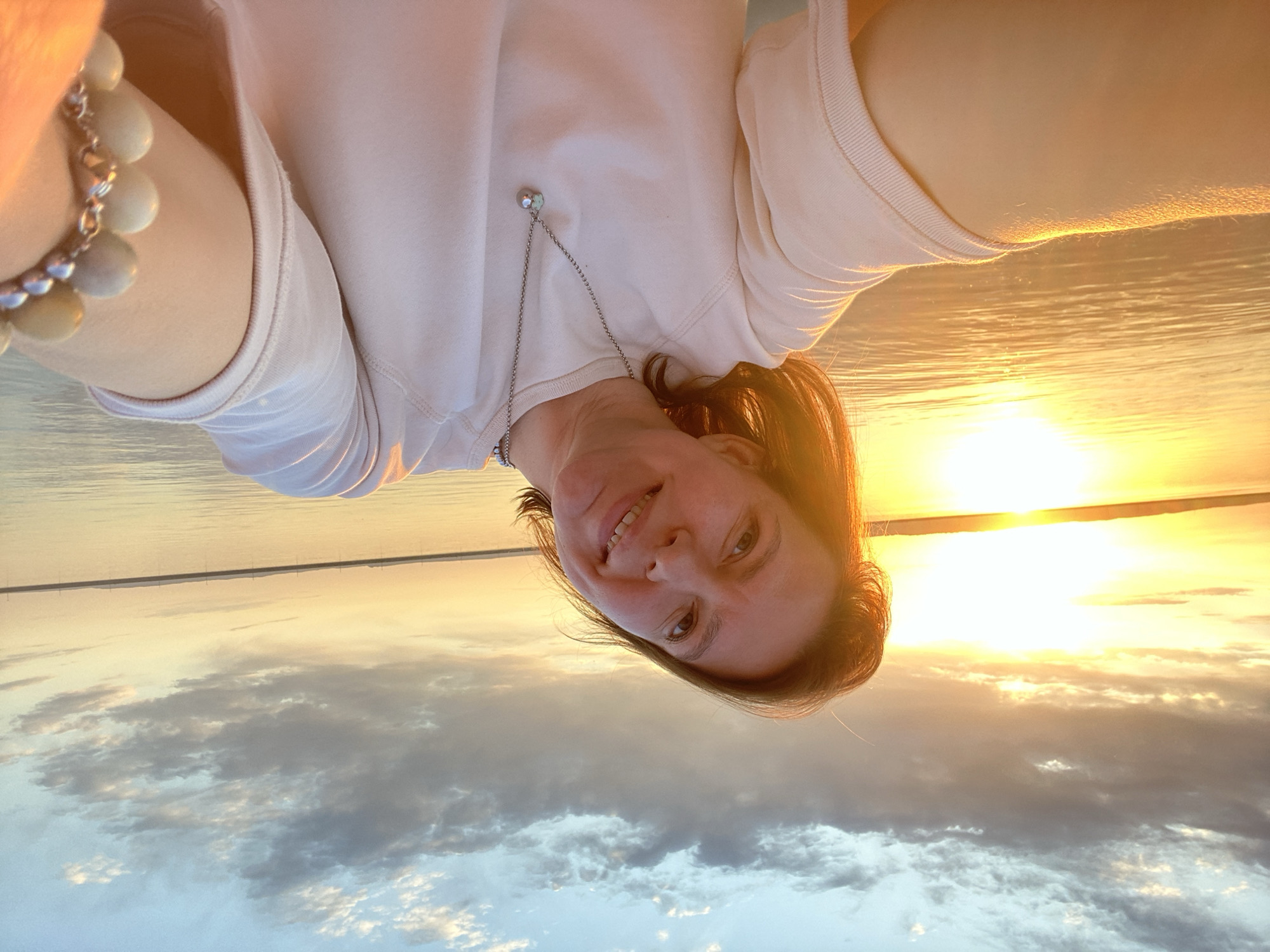 A woman is taking a selfie. She is wearing a light-colored sweatshirt and has a necklace. In the background, there is a body of water with a sunset. The photo was likely taken in the city of Chaikovsky, Russia, based on the prompt. The sky contains clouds.