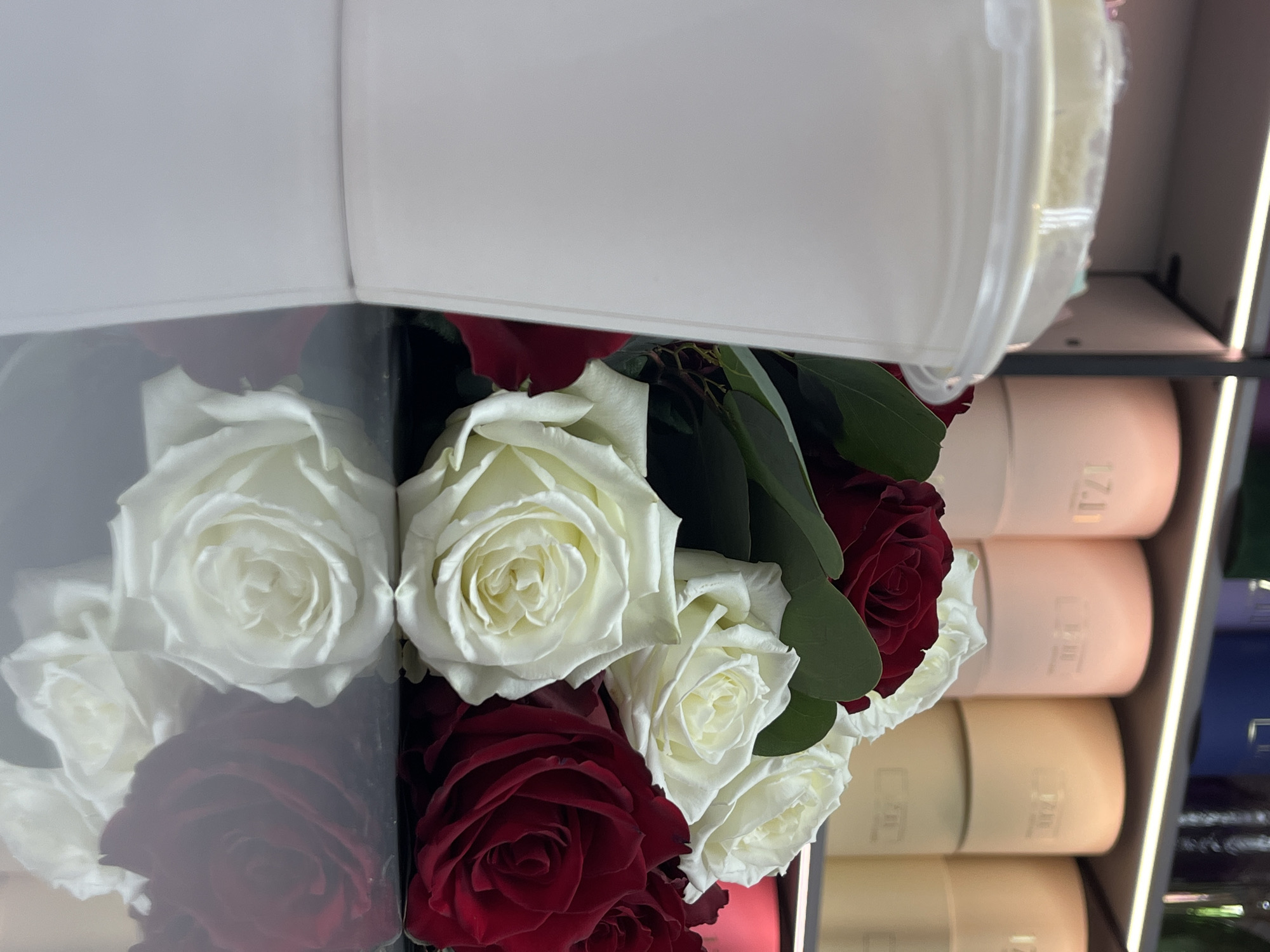 A photograph taken in what appears to be a shop in the city of Chaikovsky, Russia, features a bouquet of red and white roses in a black box. A clear plastic lid is partially obscuring the flowers. In the background are stacks of cylindrical containers in shades of tan and light pink. Vertical lighting elements are positioned to the right.