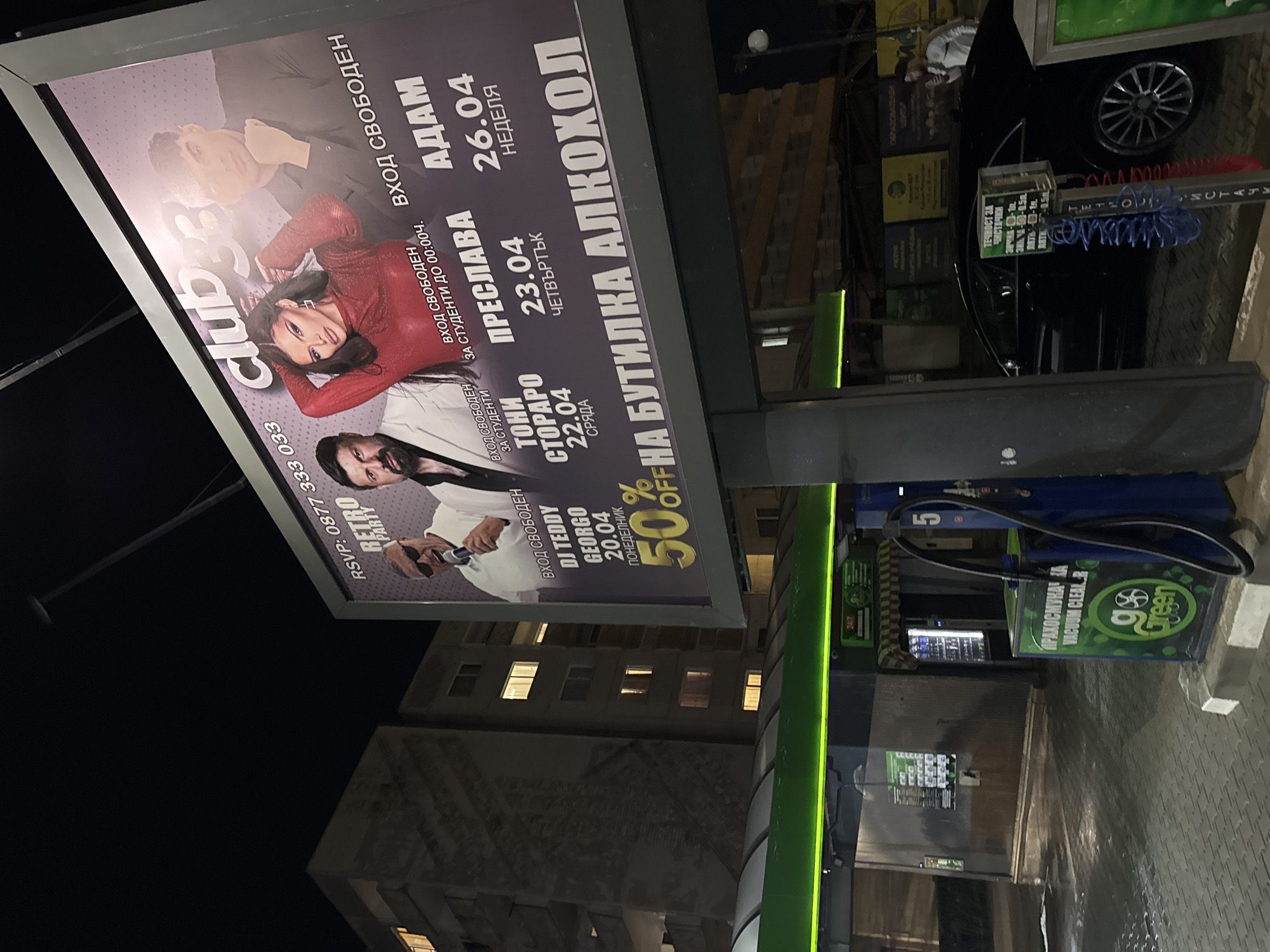 The image shows a vertical shot of a street scene at night. A billboard advertising Club 33 is prominently displayed, advertising performances by various artists including DJ Teddy Georgio on April 20, Toni Storaro on April 22, Preslava on April 23, and Adam on April 26. The advertisement mentions "free entry" and a 50% discount on alcohol bottles. The RSVP number is 0877 333 033.
The sign also states "Вход Свободен за студенти до 00:00ч." indicating free entry for students until midnight.
Next to the sign, there is a car wash/vacuuming station, with the "Go Green" vacuum cleaner machine and various signs with pricing. A black car is partially visible. A building facade with illuminated windows is visible in the background. The location is likely in Sofia, Bulgaria due to the presence of Bulgarian text on the signs.