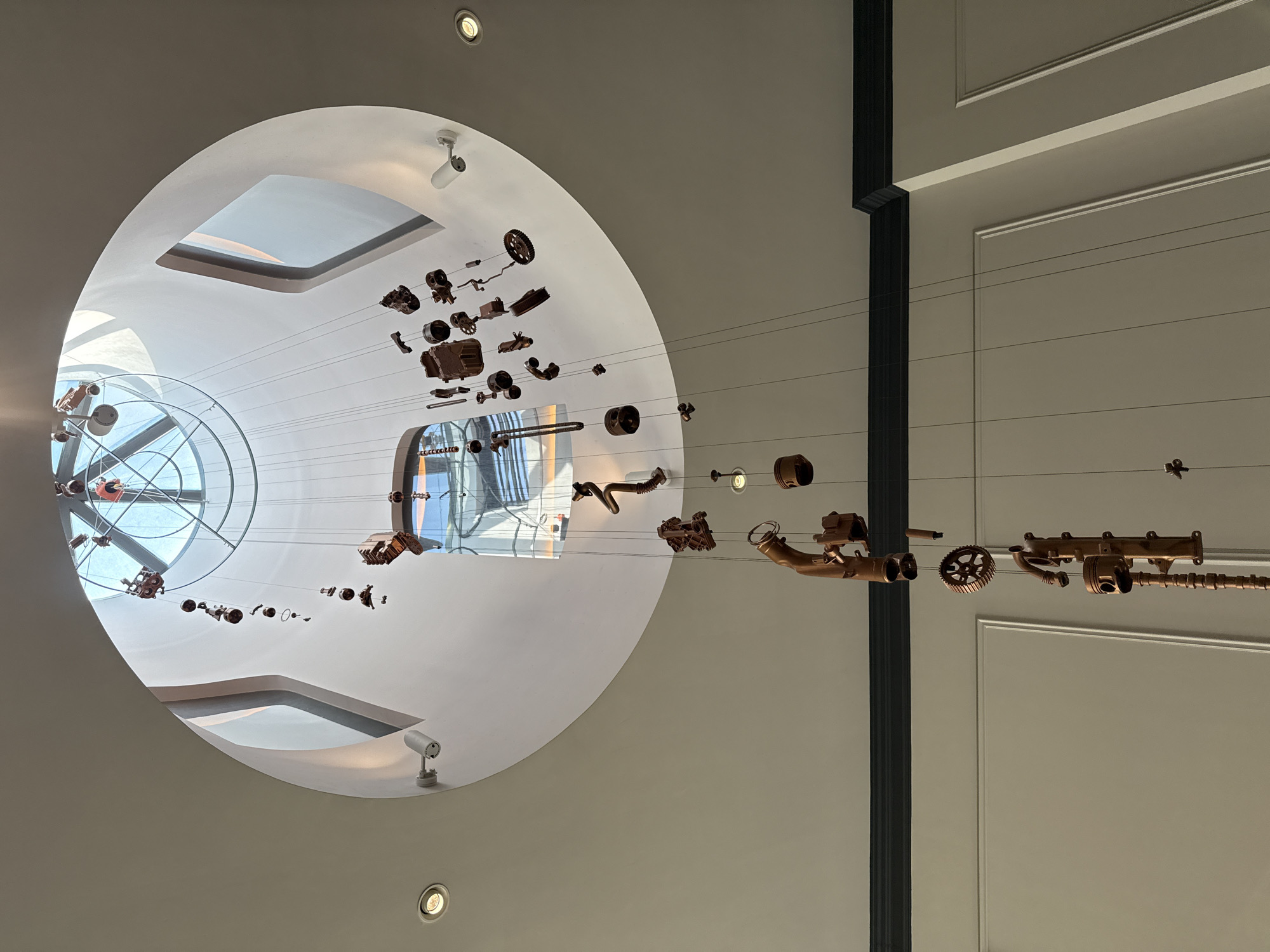 The image shows a complex art installation suspended from the ceiling inside a building. The installation appears to be composed of deconstructed machine parts, possibly engine or mechanical components, hanging on thin wires. These parts are bronze or copper-colored. The background features architectural elements including a circular skylight with visible window structures and supports beyond. Natural light is coming through this skylight. There are recessed lighting fixtures on the ceiling and a dark vertical structural beam or support is on the right. The walls are a light beige or off-white color. The building is located in Vila Verde, Portugal.