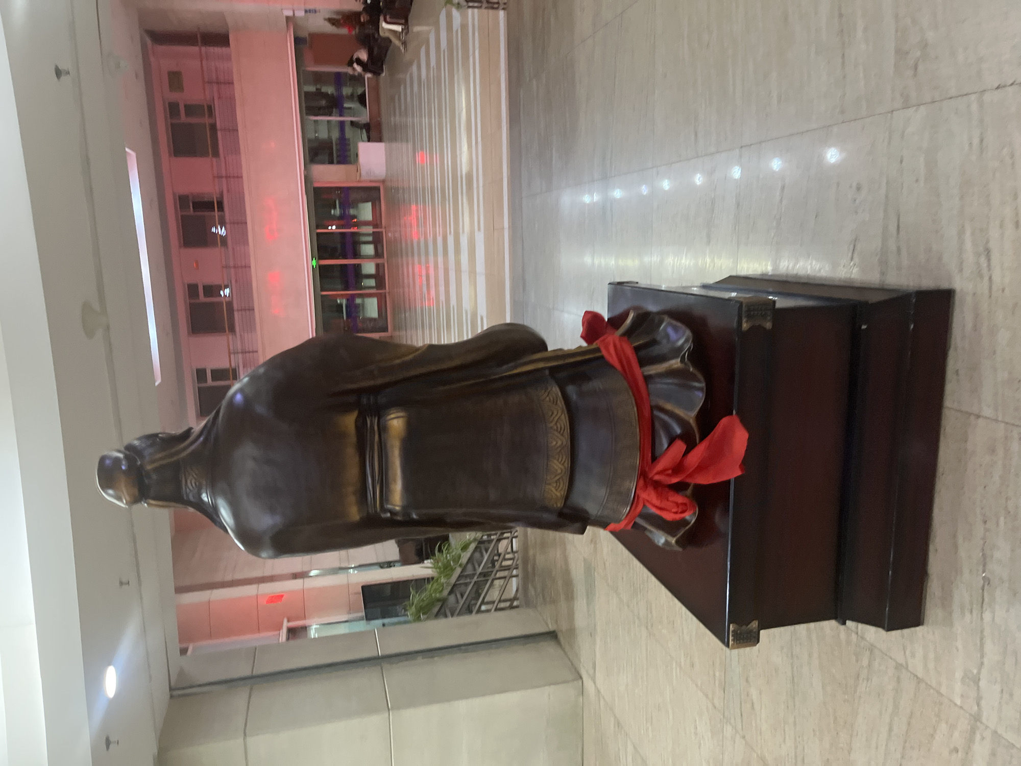 A bronze statue of a male figure, possibly a scholar or historical figure, stands on a dark wooden pedestal with a red ribbon tied around it. The statue is located in a building in Changchun City, China. The background includes a modern building with multiple floors, a tiled floor, and indoor plants. The scene is well-lit with overhead lights.