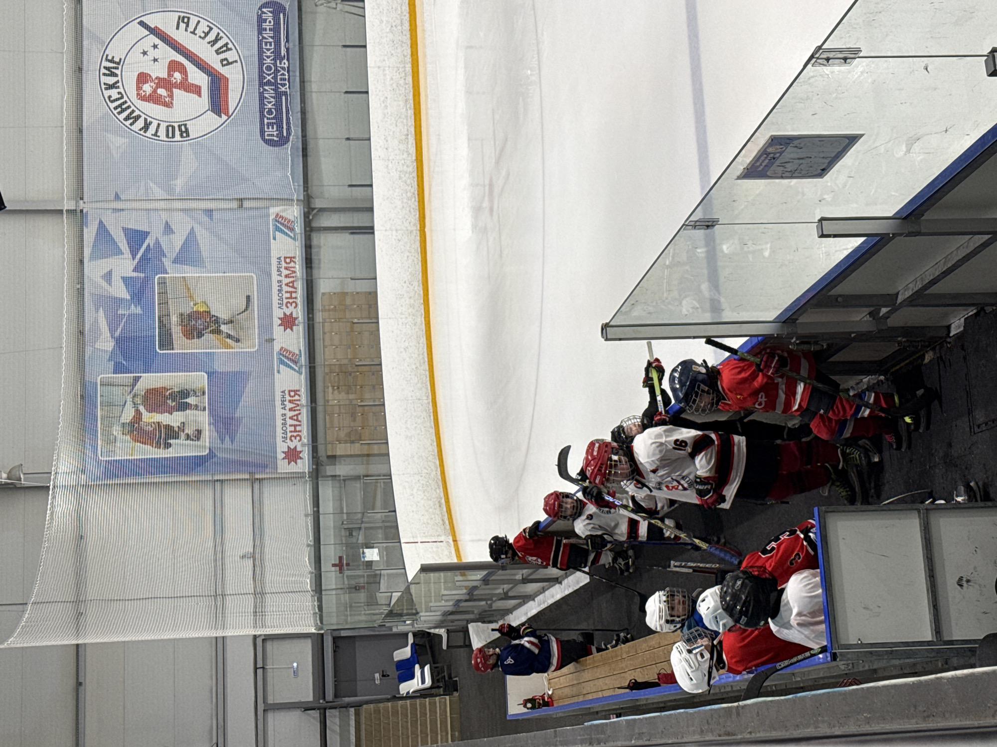 This image shows a group of young hockey players sitting on a bench at an ice rink in Votkinsk, Russia. They are wearing their hockey gear, including helmets, jerseys (red, white, and blue), and skates. The players hold hockey sticks. The ice rink is enclosed by glass barriers. A large banner displays the logo for "Votkinskiye Rakety" (Votkinsk Rockets), a children's hockey club. Another banner includes the phrase "Ledovaya Arena Znamya" (Ice Arena Banner). The image is indoors, with artificial lighting illuminating the scene.