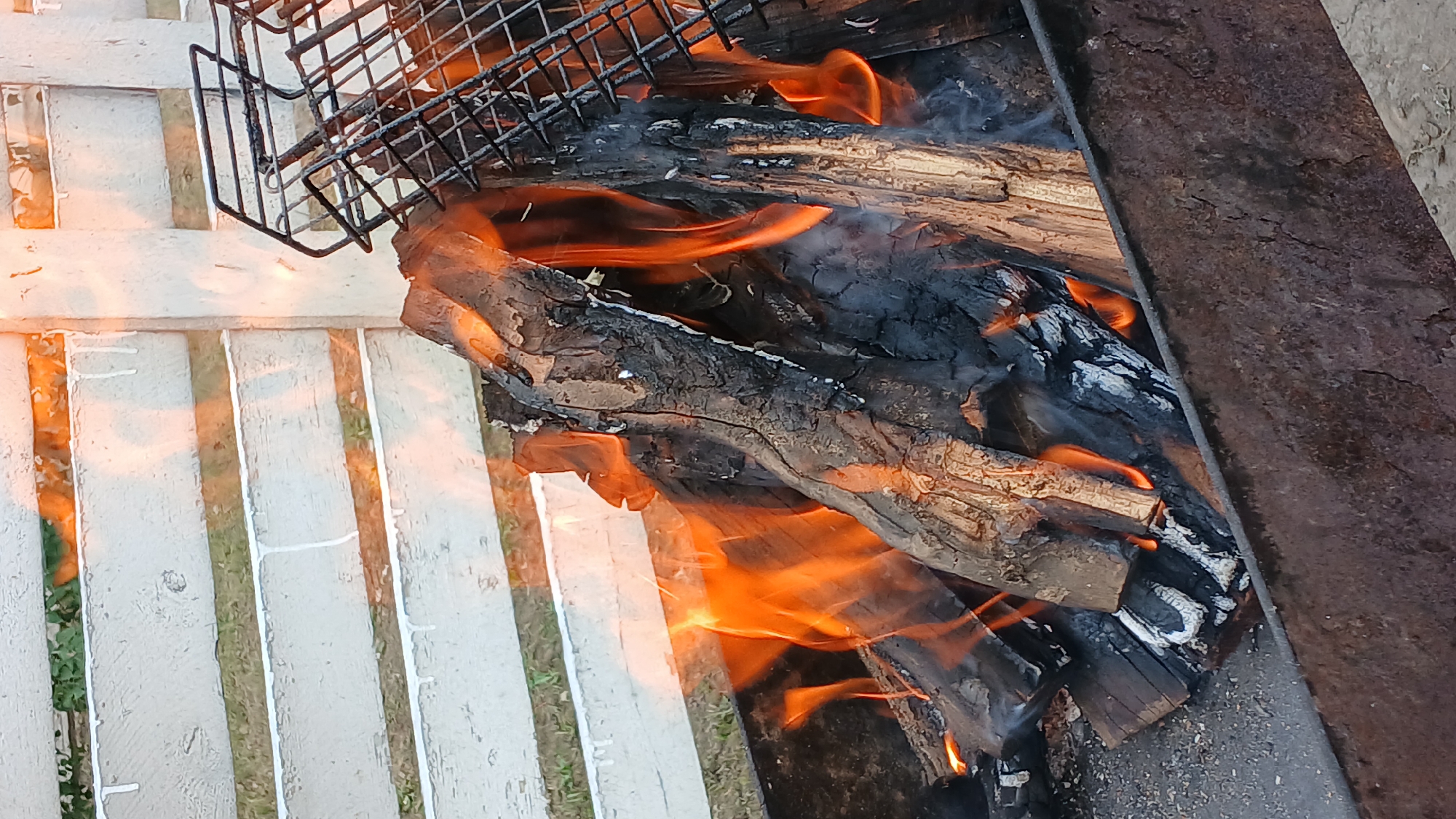 A close-up shot focuses on a fire burning in a metal brazier. Logs of wood, charred and cracked, are engulfed in vibrant orange flames. A wire grill rack sits at an angle on the right side of the frame, partially obscured by smoke and embers. In the background, a white wooden fence with horizontal slats is visible, with a hint of green foliage behind it. The lighting suggests it is daytime, with the fire's glow illuminating the immediate surroundings. There are no people or animals present in the image. The setting appears to be outdoors, likely a backyard or patio area. The location is identified as Derköl, Kazakhstan.