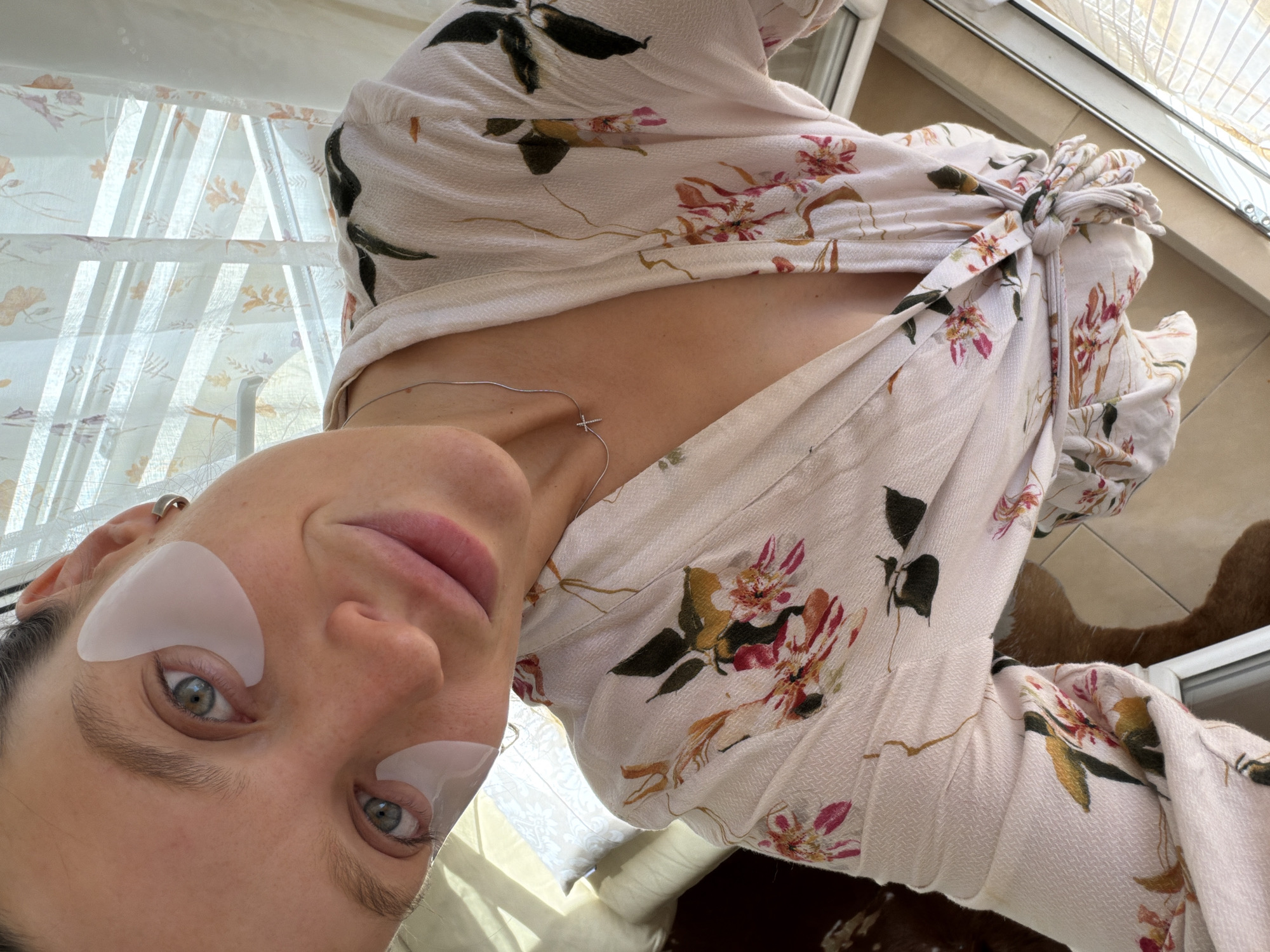 This close-up, inverted selfie captures a person, likely female, engaged in a self-care routine in an indoor setting in Budva, Montenegro. The subject has light skin, striking blue-green eyes, and a subtle, content smile. She is wearing white, gel-like eye patches beneath both eyes, indicating a beauty treatment. A delicate silver necklace with a small cross pendant is visible around her neck, resting on her chest.
She is dressed in a light-colored garment, possibly cream or light pink, adorned with a vibrant floral pattern that includes hues of pink, orange, black, and green. The clothing appears to be a loose-fitting top or dress, potentially a wrap style, with a decorative knot visible near the shoulder.
The background reveals a brightly lit indoor space, presumably near a window or glass door. Sheer, light-colored curtains, possibly with a subtle pattern, filter ample natural sunlight, casting distinct shadow lines across the scene. A white window or door frame is visible, and a soft, textured brown object, possibly an item of furniture or flooring, can be seen further in the background. The overall mood is serene and relaxed, reflecting a moment of personal care in a well-lit environment. No text is visible in the image.