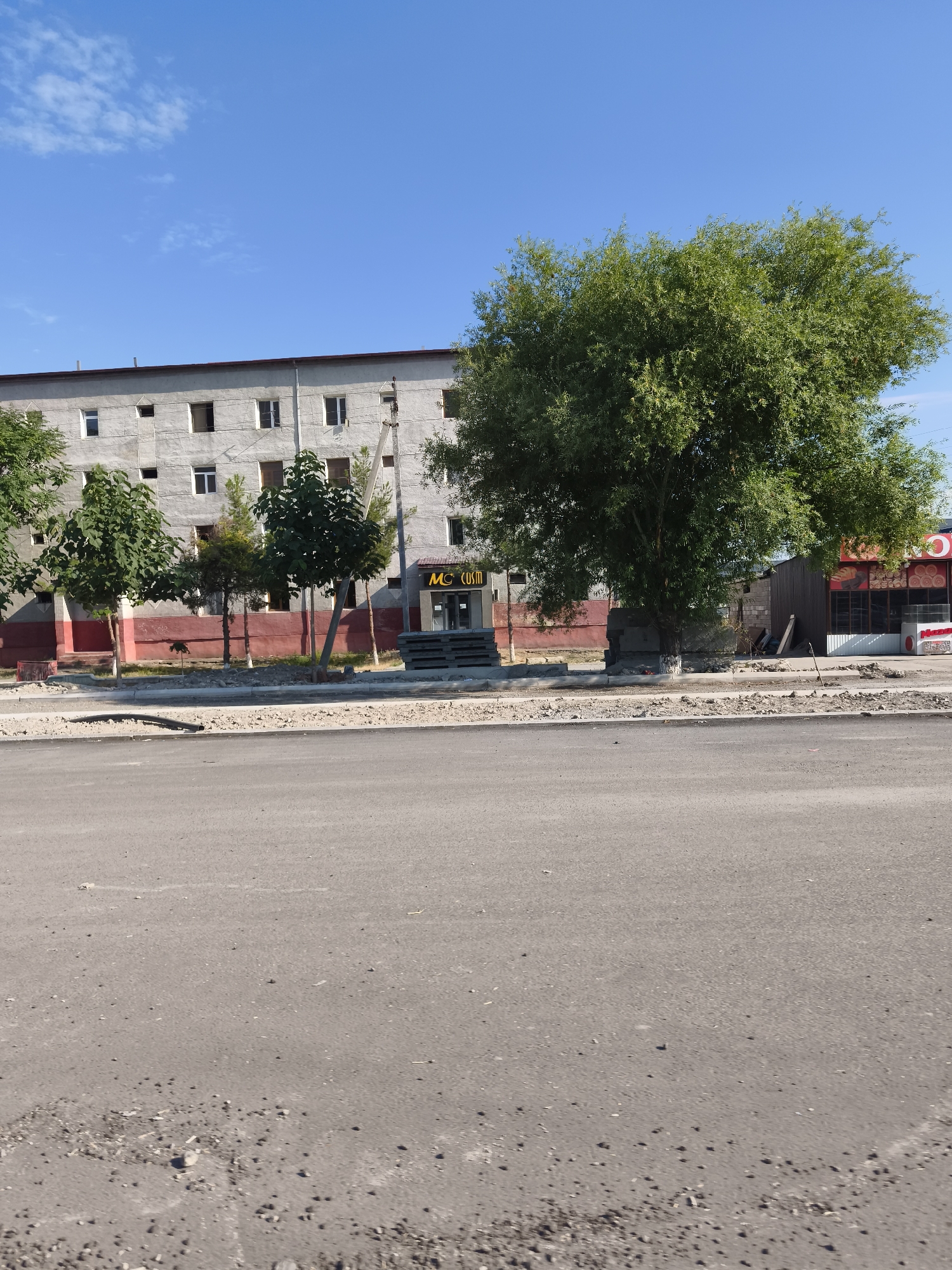 The image depicts a street scene in Buz, Uzbekistan, on a clear, sunny day. In the background, a long, multi-story building with a rough, concrete facade and a dark red lower section stands. The building has numerous windows, some open and some closed. In front of the building, a large, leafy tree dominates the right side of the frame, casting a shadow. To the left of the tree are several smaller trees and a small, booth-like structure with signage that reads "MC COSIM". Piles of what appear to be concrete blocks or construction materials are stacked near the booth and the building. The foreground shows a paved road with some debris and uneven patches, suggesting ongoing construction or repair work. The overall environment appears to be a semi-urban area, possibly with commercial or residential buildings, set against a bright blue sky.