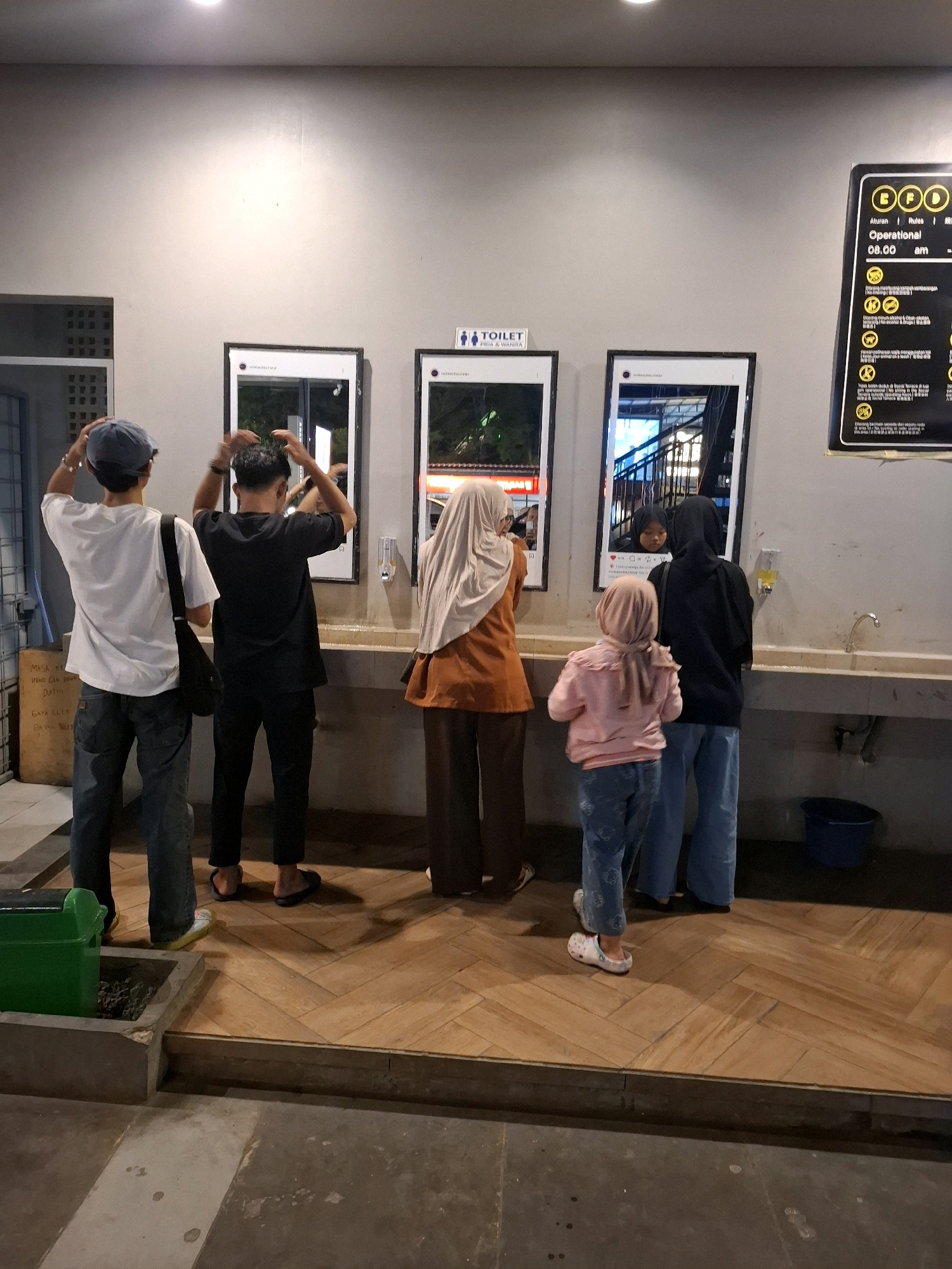 In Cianjur, Indonesia, a group of people are gathered at what appears to be a public restroom area, with several individuals interacting with mirrors mounted on the wall. Two young men are facing away from the camera, with their hands touching their heads, possibly adjusting their hair. To their right, a woman wearing a hijab and an orange top is also looking into a mirror. Further to the right, a young girl in a pink hijab and a younger child in a black hijab are standing near a sink. The mirrors are framed in a way that resembles social media posts. A sign indicating "TOILET" is visible above the leftmost mirror. To the far right, a black sign with yellow text lists operational hours and rules, indicating the facility is open from 08:00 AM. The environment features a tiled floor with a herringbone pattern and a concrete wall. A green trash bin is positioned to the left of the scene, and a blue bucket is near the sinks on the right.