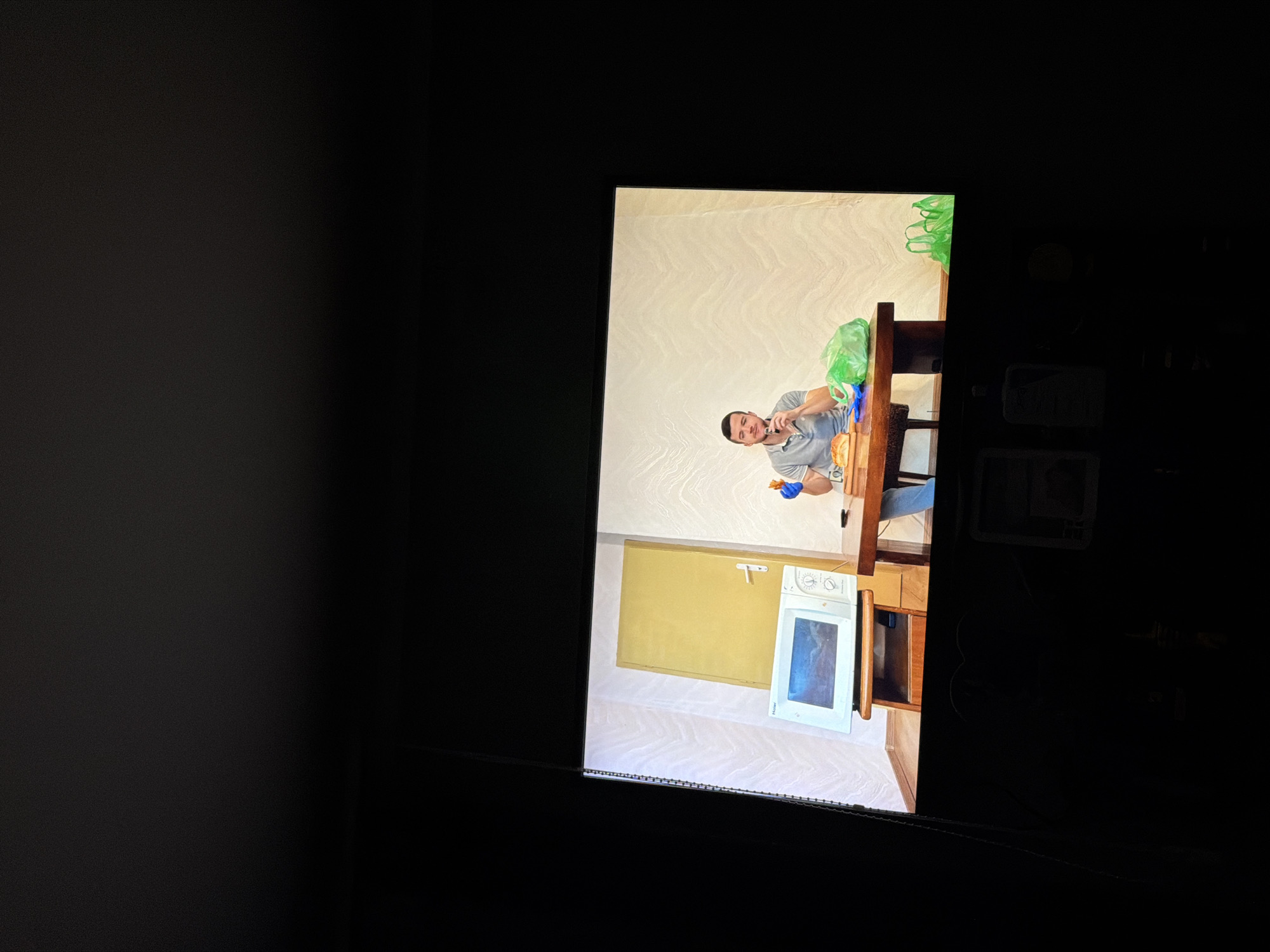 The image shows a television screen displaying a scene of a person in a kitchen. The person is wearing gloves and seems to be interacting with something on the table. There are green plastic bags on the table. A microwave and a door are visible in the background. The room where the television is located is dark. The location of the depicted scene is unknown.