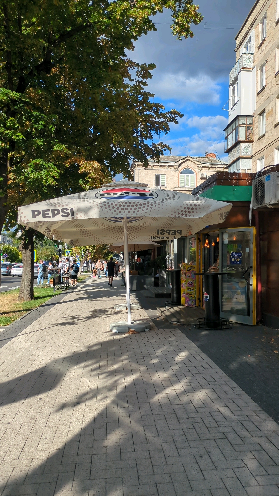 The image depicts a street scene in Chișinău, Moldova, on a sunny day with scattered clouds. A sidewalk paved with light-colored rectangular bricks runs along the right side of the frame, shaded by trees with green and yellowing leaves. A large white umbrella with the Pepsi logo is prominently featured in the foreground, extending over a portion of the sidewalk. Beyond the umbrella, a small kiosk or shop with a yellow-framed glass door is visible, offering what appears to be food and drinks, with posters on display. Several people are walking along the sidewalk and near the kiosk. Further down the street, traffic is visible, including cars and a traffic light. The background shows buildings, including a multi-story structure with balconies. The scene suggests a public outdoor area, possibly a commercial street or promenade.