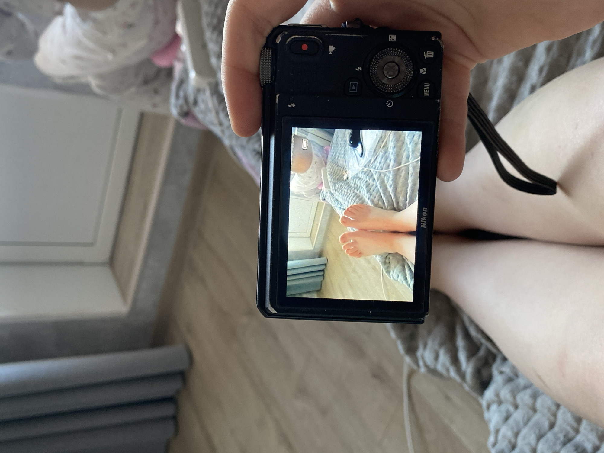 A person holds a Nikon digital camera, displaying an image on its screen. The image on the screen shows a close-up view of bare feet resting on a gray-and-white patterned bedspread. In the background of the image on the screen are a white wall and a stack of gray books. The location is Зубово, Russia.