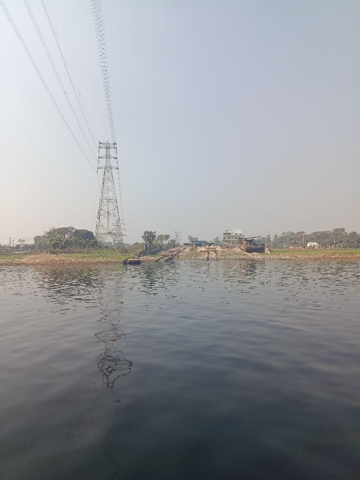 The image presents a wide, placid body of water, likely a river, under a bright and slightly hazy sky. The lower half of the frame is dominated by the dark, reflective water, exhibiting gentle ripples and a clear, distorted reflection of a tall power pylon.
On the distant bank, situated in what appears to be a semi-rural or industrial area within Zazira, Bangladesh, a prominent metal lattice power transmission tower stands on the left. Multiple power lines extend from the pylon and across the clear sky. The shoreline is rugged, featuring patches of exposed earth, scattered green vegetation including trees and shrubs, and several man-made structures.
To the right of the power pylon, a cluster of buildings is visible. A notable multi-story structure with a white upper section and a darker base stands out, featuring a sign on its facade that clearly displays "MOHAMMAD SHARIF" and "BANGLADESH" among other indistinct text. Adjacent to these buildings, what appear to be ramps or slipways extend from the bank into the water, suggesting their use for boats or transport. Further along the bank to the right, smaller, simpler structures are scattered amidst the sparse vegetation.
The scene is static, with no discernible human figures or active events taking place. The time of day is daytime, likely morning or afternoon, characterized by the even, clear lighting and the absence of strong shadows. The weather is clear, though a slight atmospheric haze is present. The image conveys a sense of tranquility, combining natural elements with utility infrastructure and modest development along a waterway.