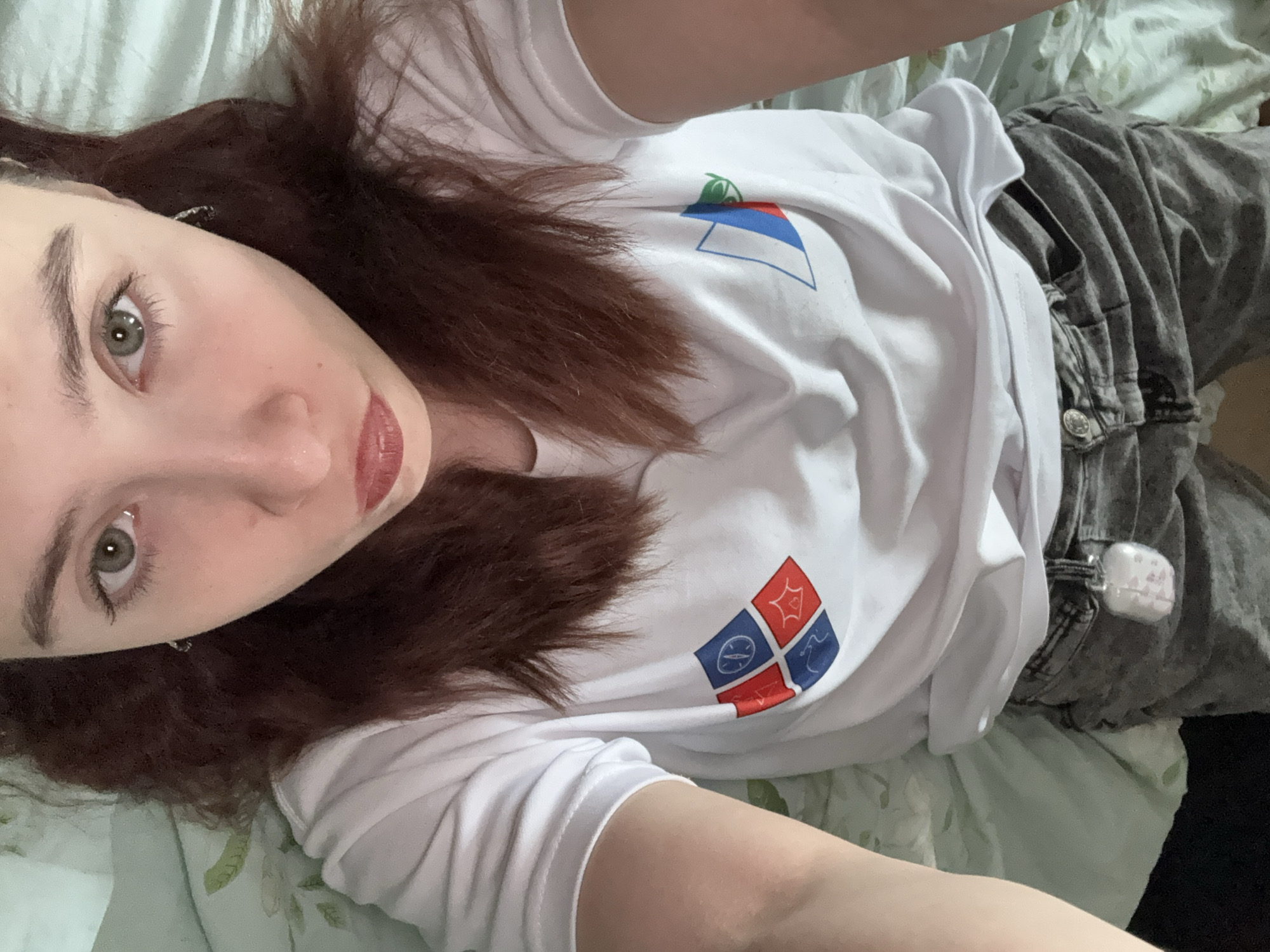 A selfie taken indoors shows a woman with shoulder-length brown hair lying on her back on a bed covered with a light green floral-patterned bedsheet. She has fair skin, grey eyes, a nose piercing, and is wearing red lipstick. She is wearing a white t-shirt with a design depicting two flags: one is a white, blue, and red tricolor, the other a square divided into four smaller squares, each containing a different symbol. She is wearing grey jeans. An object, likely a container, is hanging from one of the belt loops on her jeans. Based on the flags visible on her shirt, it is presumed this image may be taken in Berezniki, Russia.