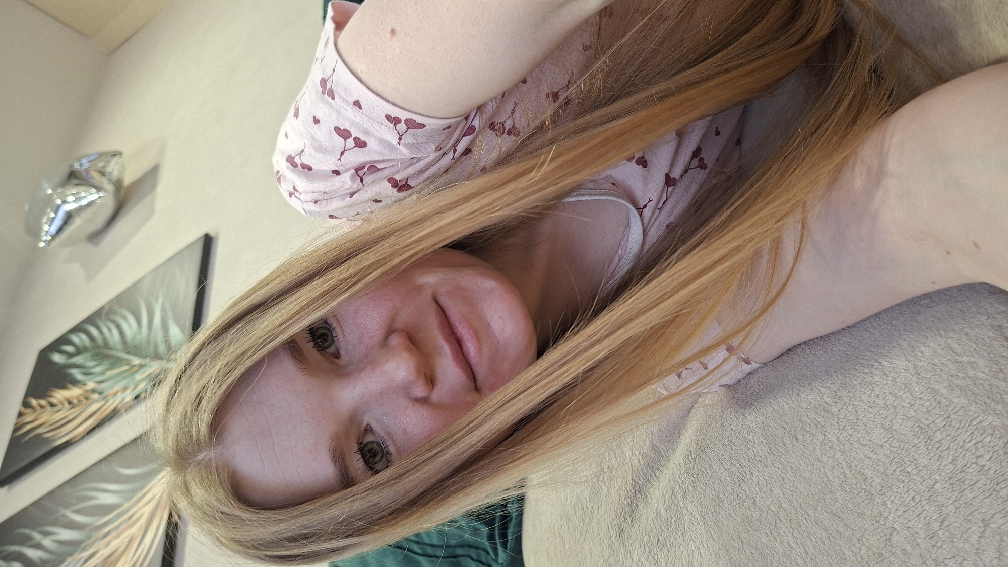 The image shows a woman lying on a light grey-colored blanket. She has blonde hair that is partially covering her face, fair skin, and green eyes. She is wearing a light pink top with a red leafy pattern. The background includes a wall with two framed pictures of green leaves and a silver star-shaped balloon. The location is in an interior space, likely a home, but the city of Chaikovsky, Russia, is not evident in the image.