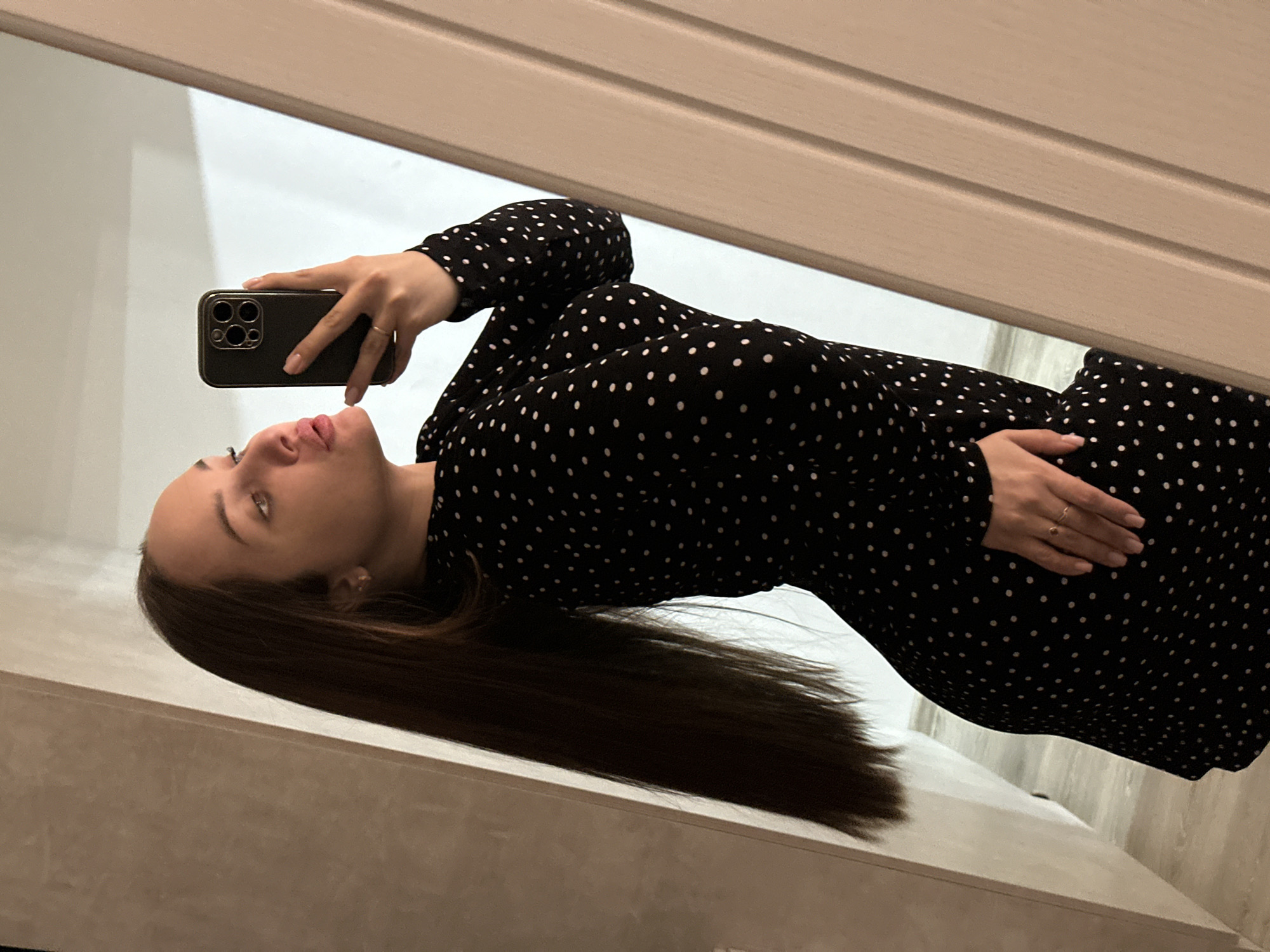 A woman is taking a selfie in Chaikovsky, Russia. She is wearing a black dress with white polka dots. Her long brown hair hangs down as she holds a black smartphone. Her right hand rests on her waist. She is standing indoors near a white surface and a light-colored wall.