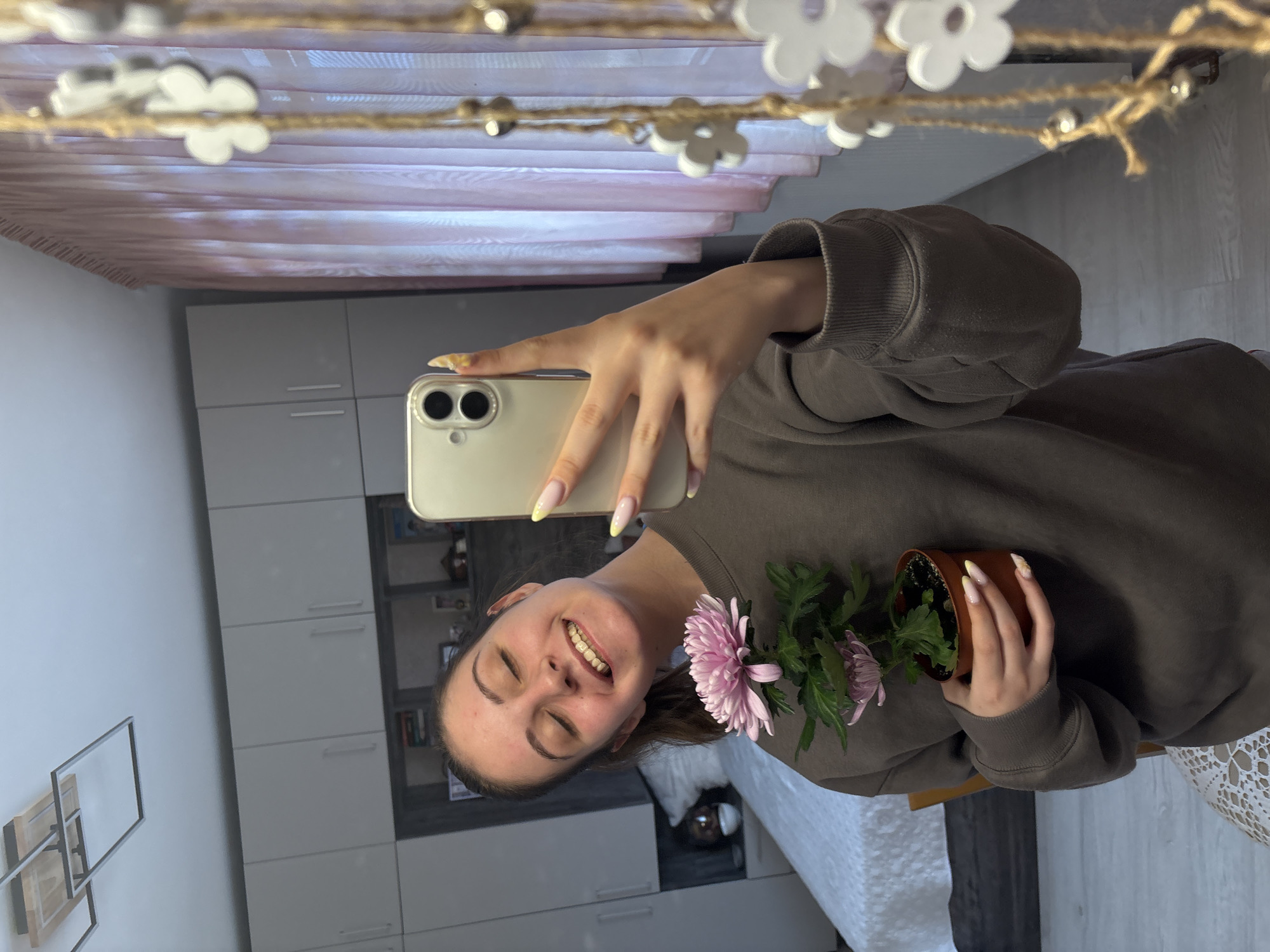 The image shows a young woman taking a selfie in a mirror. She is holding a potted pink chrysanthemum. The background reflects a modern interior with light gray cabinets and a decorative flower garland hanging across the top of the mirror. The location in Bulgarovo, Bulgaria is not verifiable from the image.