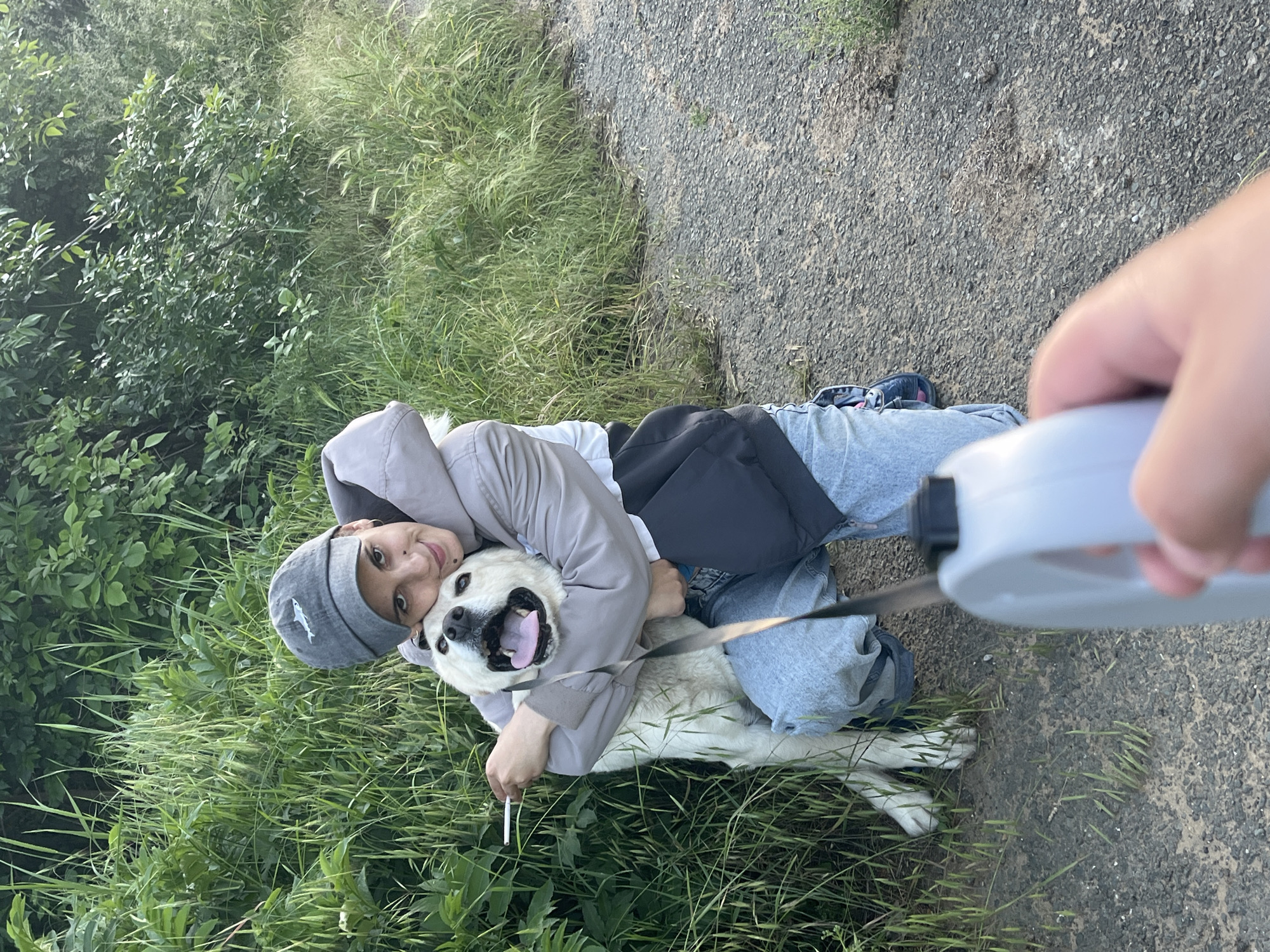 A young person kneels in a grassy area next to a paved path, embracing a large, cream-colored dog. The person wears a grey jacket, light-wash jeans, and a grey baseball cap. The dog has its mouth open. A retractable leash extends from the dog's collar, held by a hand in the foreground. The location is undetermined, it could not be determined from the visual information. There is a cigarette on the ground near the dog.