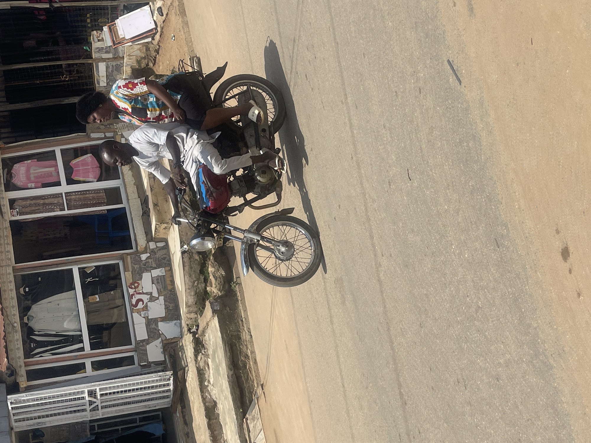 A man and a woman are riding on a motorcycle in a street in Abuja, Nigeria. The man is driving the motorcycle, and the woman is sitting behind him. The man is wearing a white shirt and grey pants, and the woman is wearing a colorful shirt and black pants. They are both wearing sandals. The motorcycle is red and appears to be a relatively old model. In the background, there is a building with a storefront. The storefront has windows displaying clothing. The street is paved with asphalt. The sun is shining, casting shadows on the road. The overall scene appears to be a typical street in an urban area.