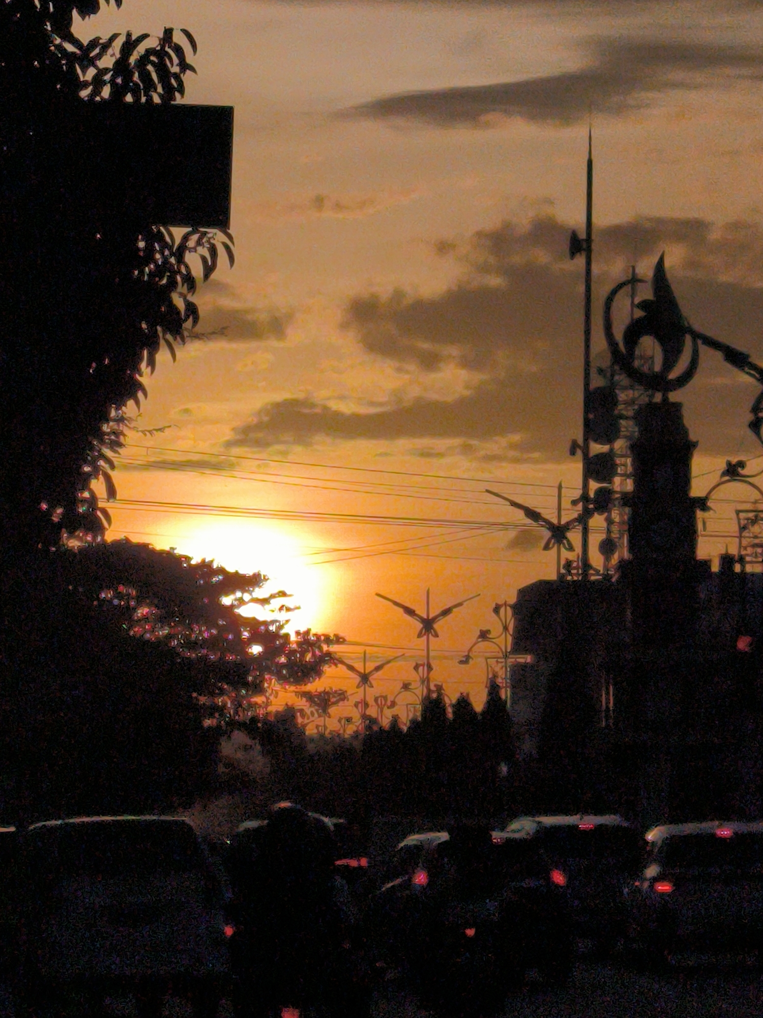 The image shows a sunset scene in Lhokseumawe, Indonesia. In the foreground, silhouetted vehicles are lined up on a road. A tree with thick foliage is on the left side, and a sign is visible among the leaves. In the background, silhouetted structures including a tower with antennas and decorative elements stand against the bright sky. Multiple bird-shaped decorations or possibly anti-bird spikes are installed along the buildings. Electrical wires stretch across the sky above the structures. The sky is a mix of yellow, orange, and cloudy grey hues.