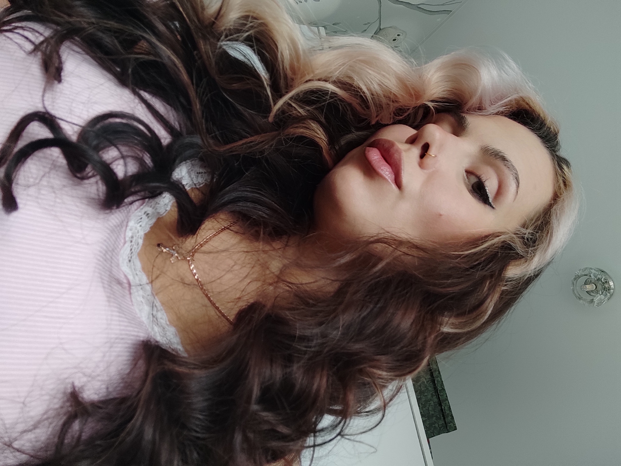 The image is a close-up, portrait-style photo of a woman with wavy hair. Her hair is dyed in a contrasting style, with dark brown dominating and sections of platinum blonde framing her face. She has fair skin, dark eyes with eyeliner, and a nose ring. She's wearing a light pink top with white lace trim and a gold necklace. The background appears to be a room interior with light-colored walls. The photo is rotated, giving a slightly disorienting perspective. There is no information in the image to suggest the city of Chaikovsky, Russia.