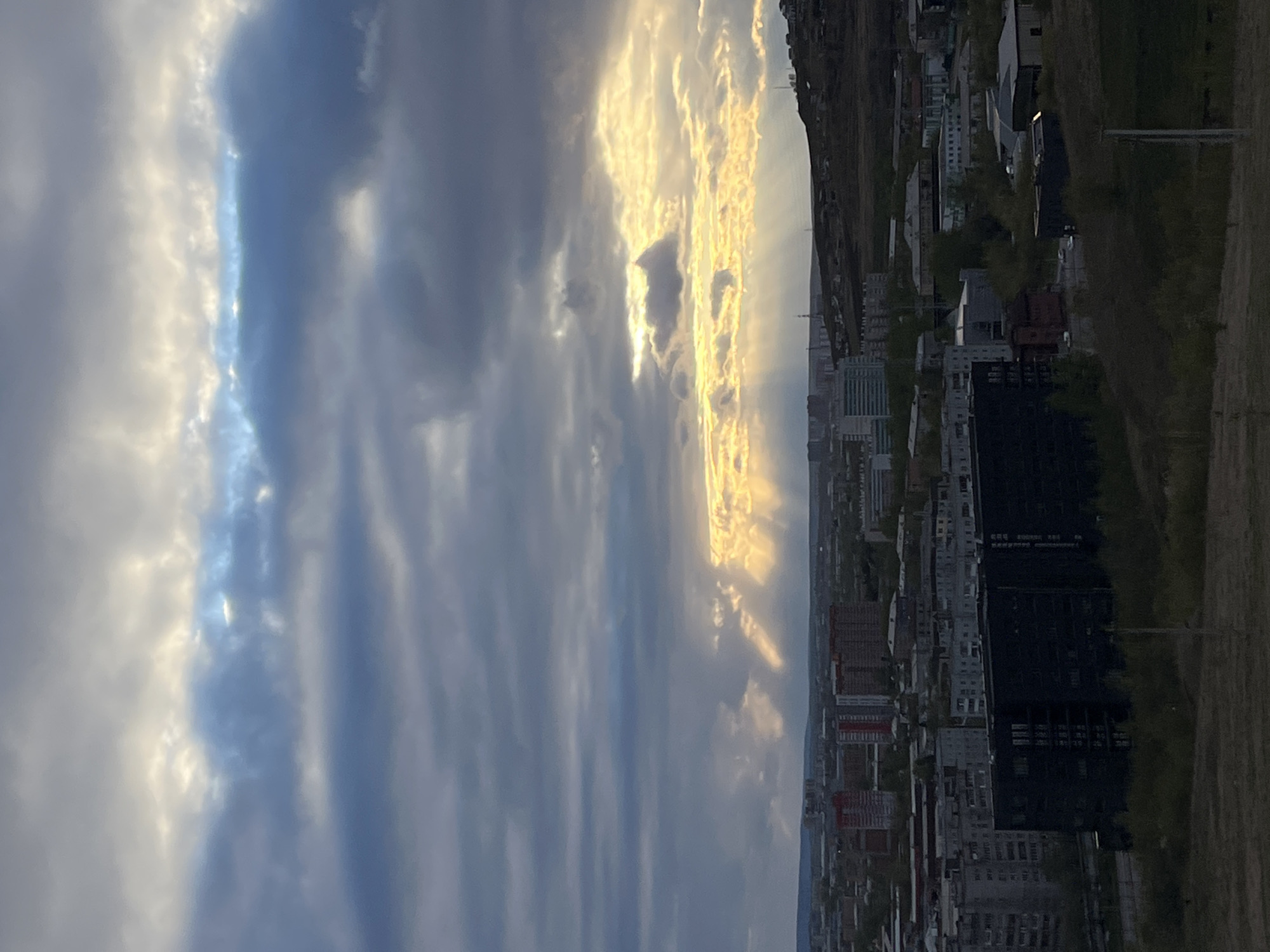 The image depicts a cityscape in Krasnoyarsk, Russia, under a dramatic sky. The sky is filled with layered clouds, illuminated by the setting or rising sun, creating rays of light. On the right side of the frame, there is a view of a built environment, including residential and commercial buildings, with a dark, tall building dominating the skyline. A natural landscape, possibly a hillside covered in vegetation, borders the city. The scene appears to be captured from an elevated vantage point, providing a panoramic view of the cityscape and the sky above.
