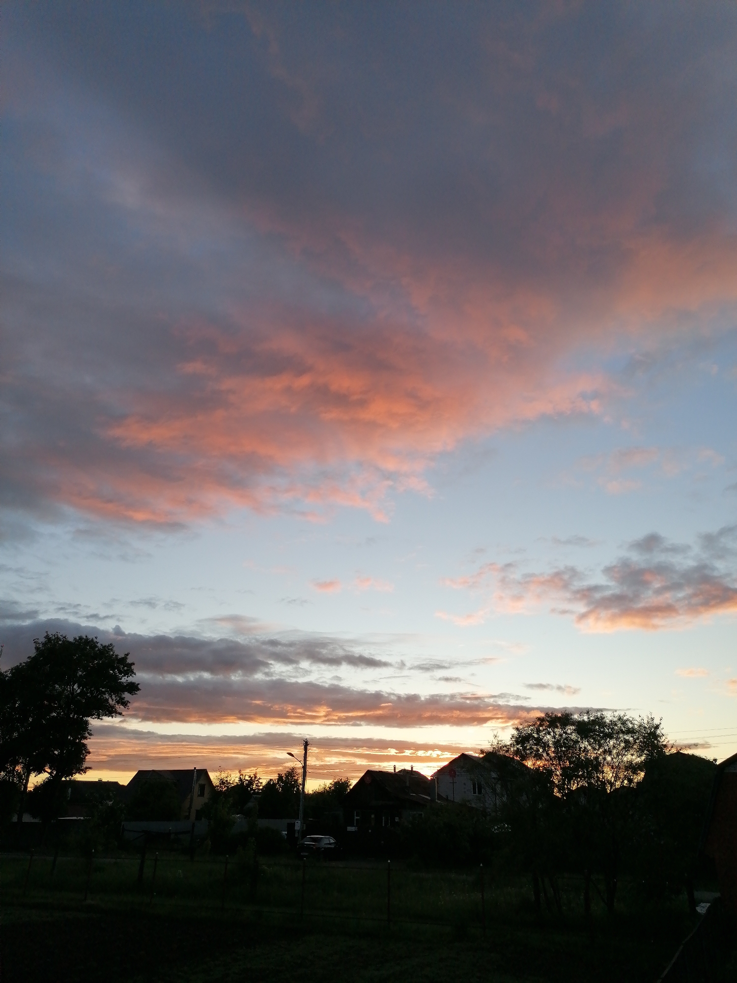 The image depicts a twilight scene in a likely rural or suburban setting. The sky is dominated by pink and purple-hued clouds, with patches of blue visible. Below the horizon line, silhouettes of houses, trees, and a utility pole are visible. A car is parked on a street. The foreground consists of dark vegetation. The location is Unknown, Unknown.