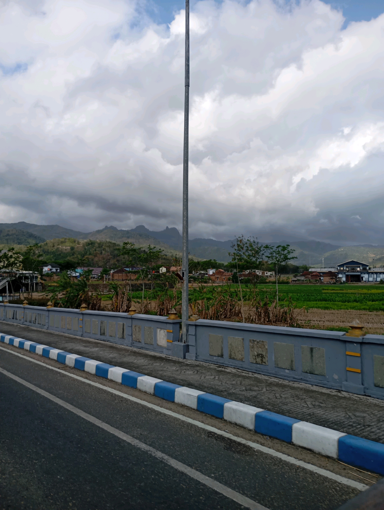 The image shows a view from a bridge in Pacitan, Indonesia, overlooking a rural landscape. A gray concrete bridge railing with decorative rectangular insets runs along the right side of the frame. A road with a white line runs in the foreground. Beyond the bridge, a village is visible with houses and buildings, nestled among green fields and trees. In the background, rolling hills and mountains are shrouded in dramatic, cloudy skies. A tall, slender lamppost stands prominently on the bridge. The scene appears to be overcast and potentially humid.