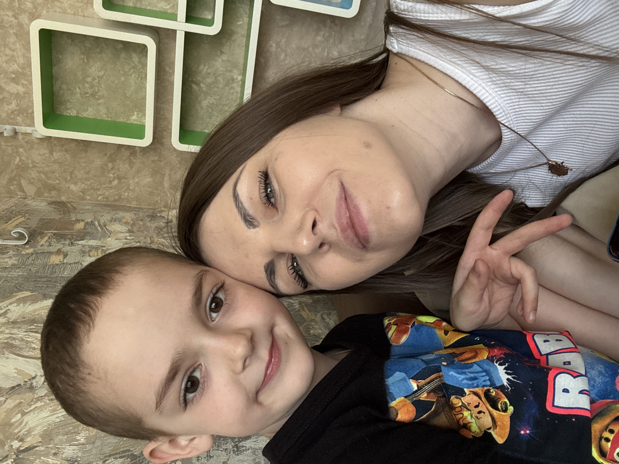 Two people, an adult female and a young boy, are posing for a selfie-style photograph. The woman has long brown hair, fair skin, and is wearing a white striped shirt and a necklace. She is holding up a peace sign with her right hand. The young boy has short brown hair and is wearing a black shirt with a colorful graphic. The background consists of patterned walls with square decorations. The location is Razdolye, Russia.