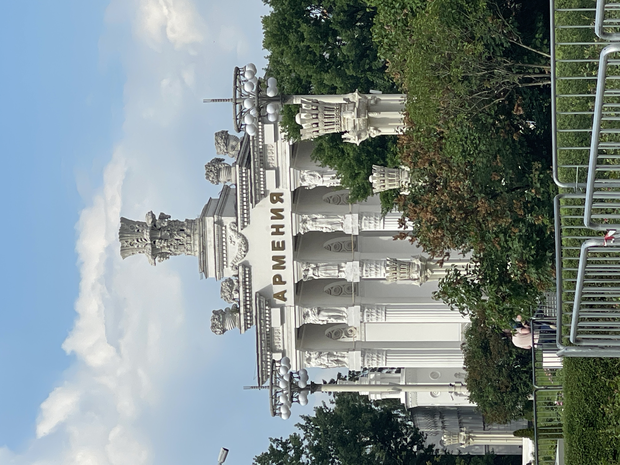 A sunlit, outdoor shot captures a grand, white architectural structure with the word "АРМЕНИЯ" emblazoned in gold lettering above a colonnade of statues. Flanking the entrance are ornate lampposts with multiple light fixtures and elaborate decorative columns. Lush green trees and foliage surround the structure, with a bright blue sky and scattered white clouds above. In the foreground, a metal fence separates the viewer from a manicured hedge and a small area with two people seated on a bench. The scene appears to be a public space, possibly an exhibition or park, given the architectural style and surrounding landscaping.