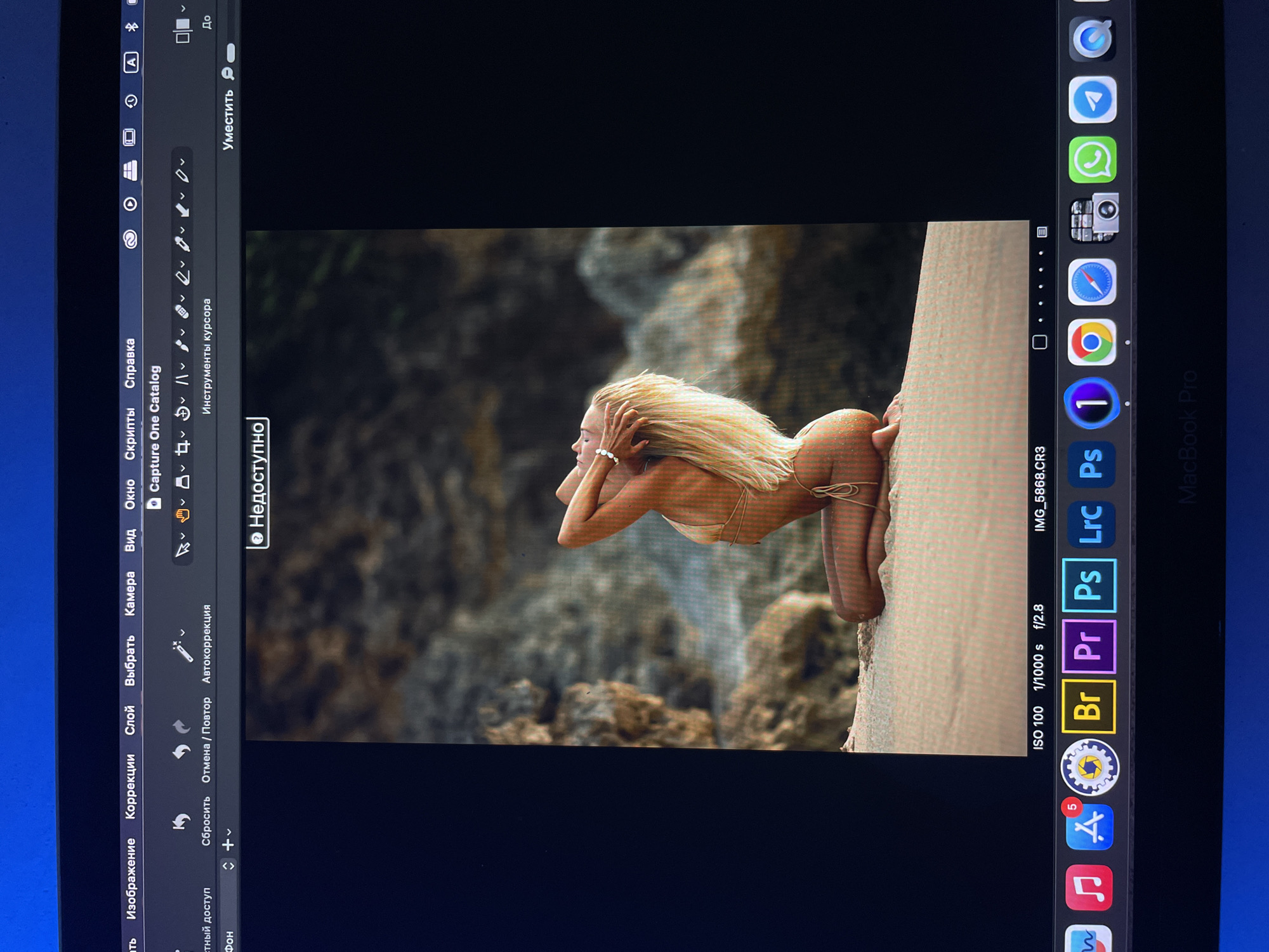 The image is a photograph of a woman kneeling on a rock formation, likely a cliff face, with a rocky background. She is wearing a skimpy, shiny swimsuit. Her blonde hair is styled upwards, and she has her hands near her head. The photo appears to be displayed on a MacBook Pro screen. The screen also shows the interface of an image editing software, possibly Capture One, with various tools and menus visible. The Dock at the bottom of the screen shows icons for applications like Final Cut Pro, Adobe Lightroom, Photoshop, Adobe Premiere Pro, Google Chrome, Safari, and others. The filename of the image is IMG_5868.CR3. The image's EXIF data indicates ISO 100, 1/1000 s shutter speed, and f/2.8 aperture. There is no information about Kazan or Russia in the image.